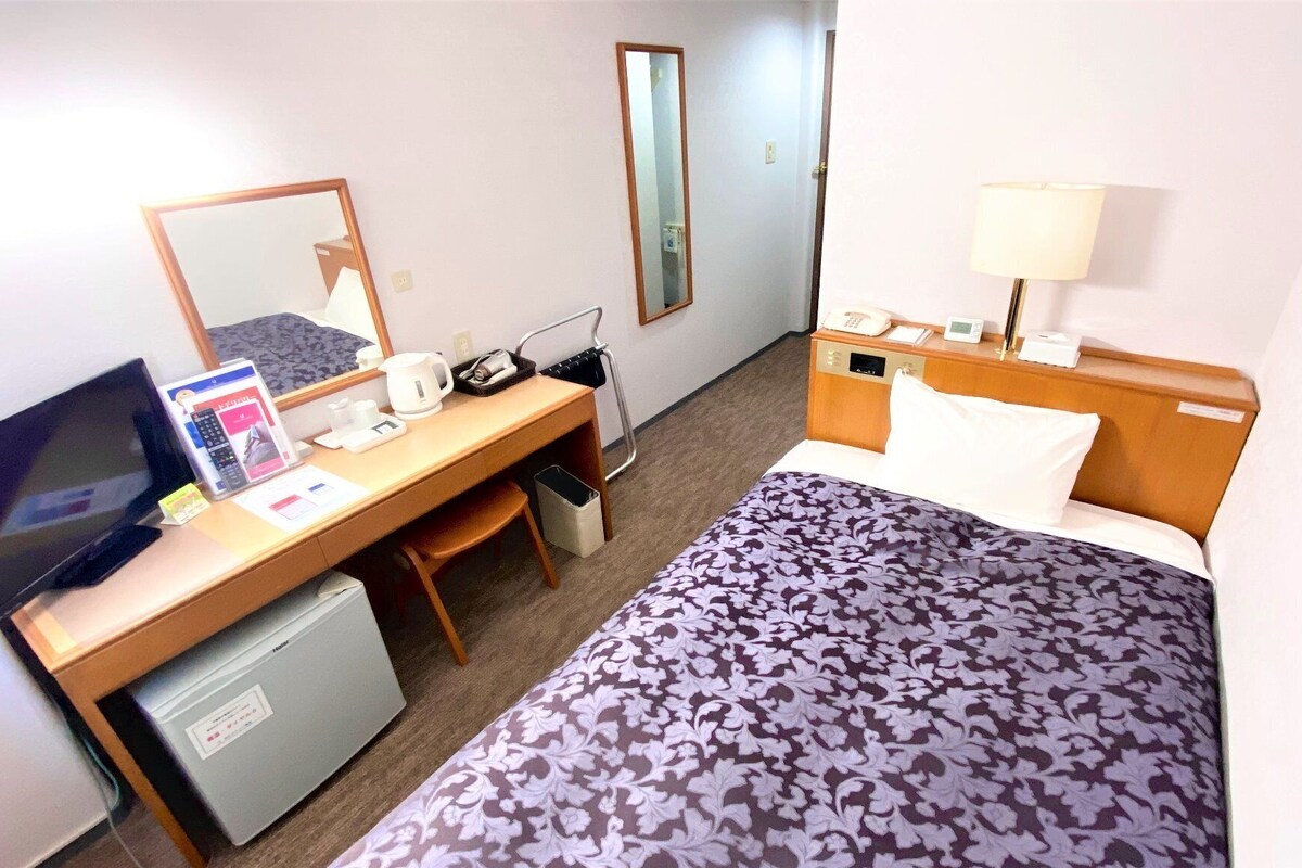 A hotel room with a bed, a desk with a computer, a television, a mirror, a lamp, and a small refrigerator.