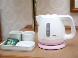 A white and pink electric kettle with a tea pod holder, two white cups, and a green tea packet on a wooden surface. - Hotel New Saitama (Saitama)