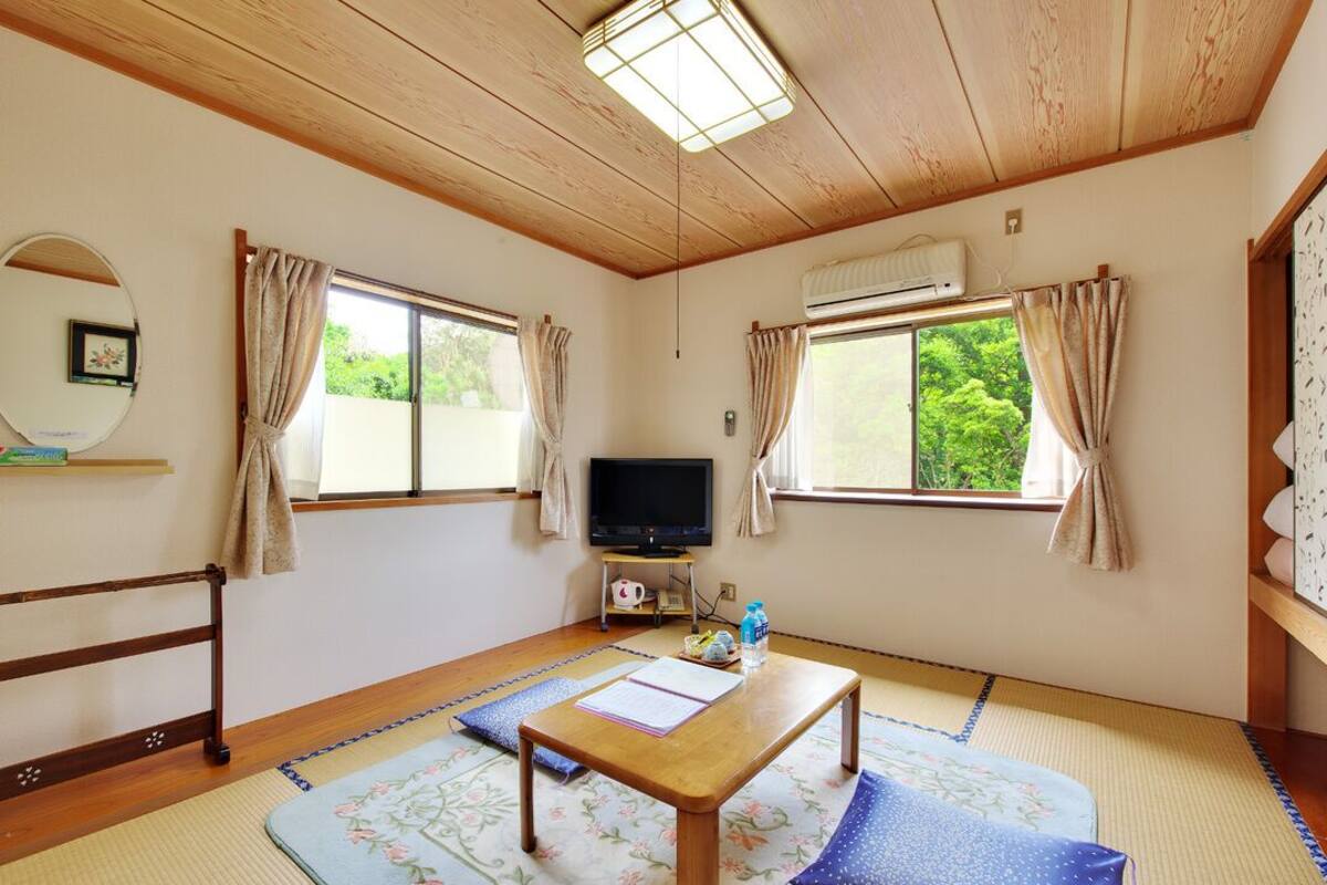 A room with tatami flooring, a wooden table, a television, and a view of greenery through the windows.