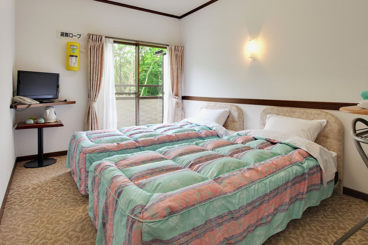 A hotel room with a bed, a television on a stand, a small table with a kettle, and a window with curtains.