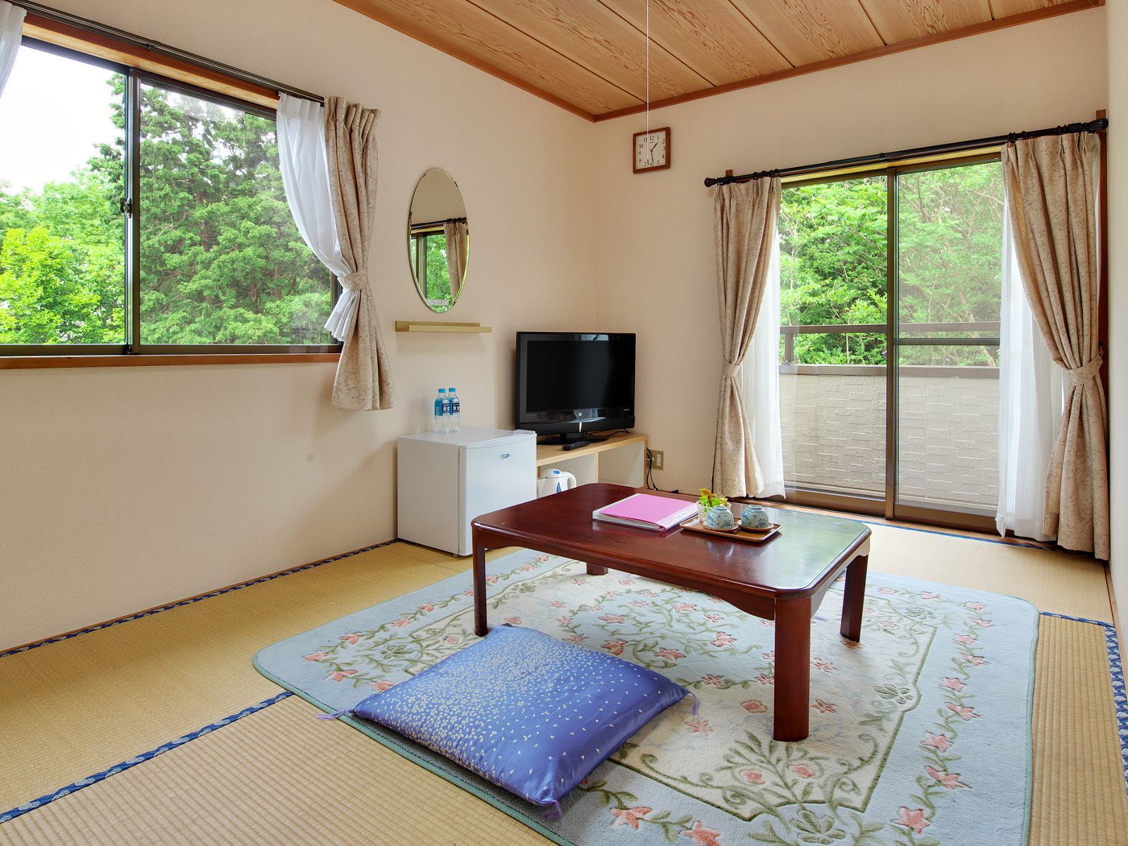 A room with tatami mats, a wooden table, a television, a small refrigerator, and large windows with curtains.