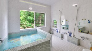 A modern bathroom with a large tiled bathtub, a window with a view of greenery, a shower area, and a vanity with a mirror and sink. - Château Le Fleur (Ito)