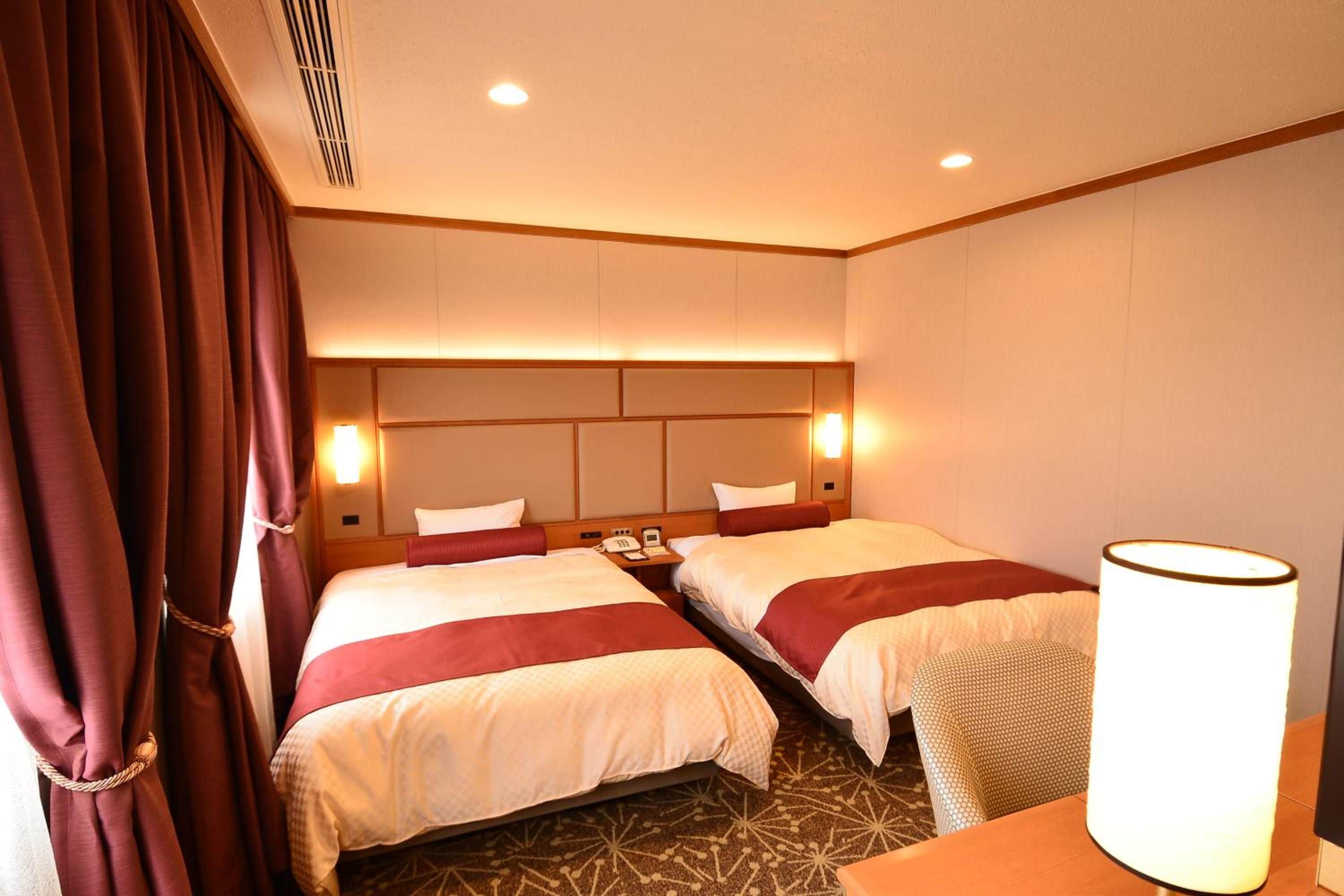 Quarto de hotel com duas camas, mesa de cabeceira, luminária e janela com cortinas.