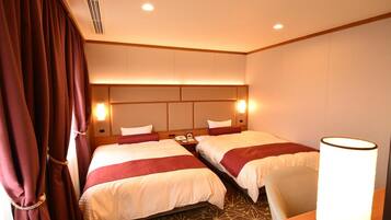 Quarto de hotel com duas camas, mesa de cabeceira, luminária e janela com cortinas.
