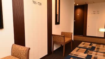 Ein Hotelkorridor mit Teppichmuster, zwei Holzsesseln und einer Wand mit Zimmernummern und einem Spiegel.