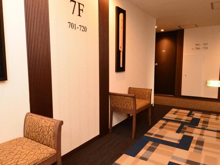 A hotel corridor with a patterned carpet, two wooden chairs, and a wall with room numbers and a mirror.