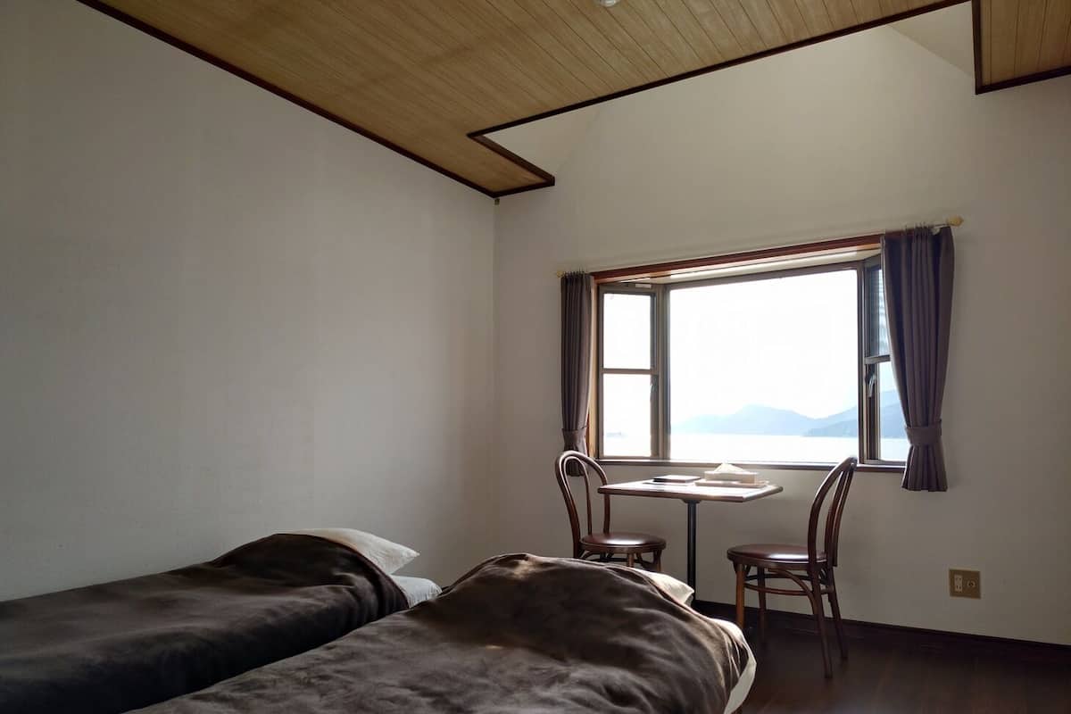 Quarto com duas camas, uma mesa pequena, uma cadeira e uma janela com cortinas.