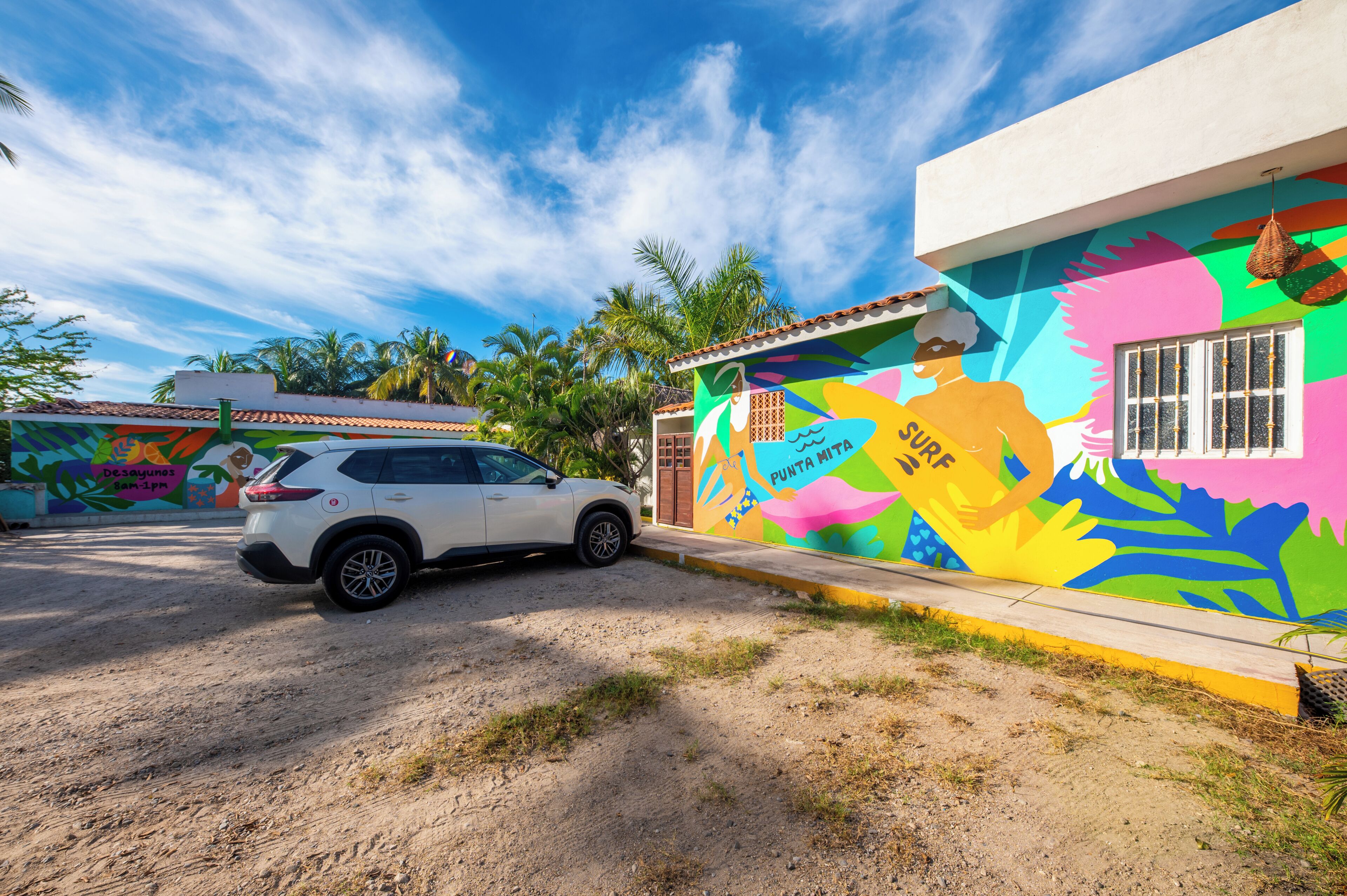 Un SUV blanco estacionado frente a un edificio colorido con un mural, palmeras y cielo despejado.