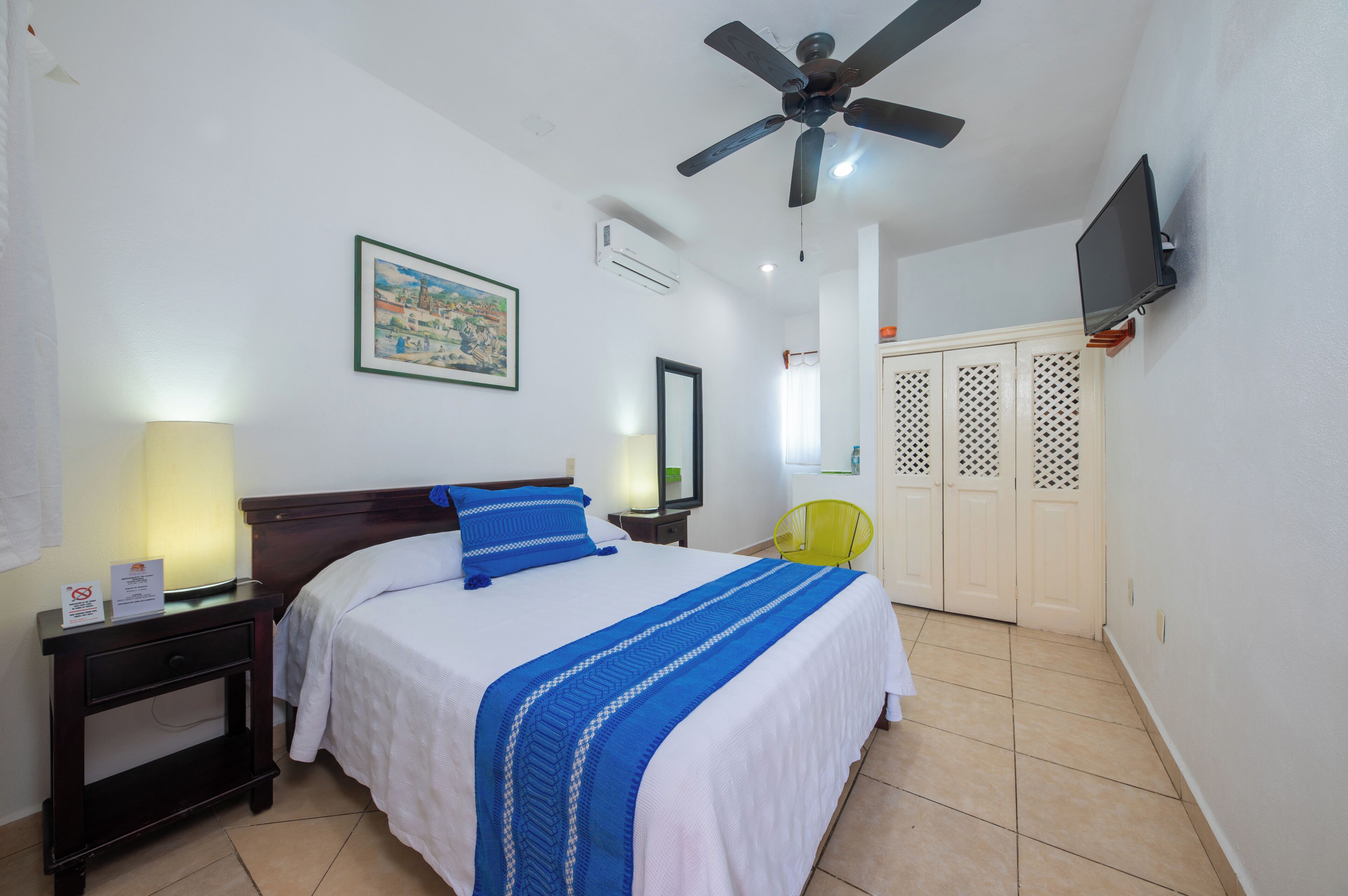 A hotel room with a bed, a nightstand, a lamp, a ceiling fan, a TV, a painting, a mirror, a chair, and a wardrobe.