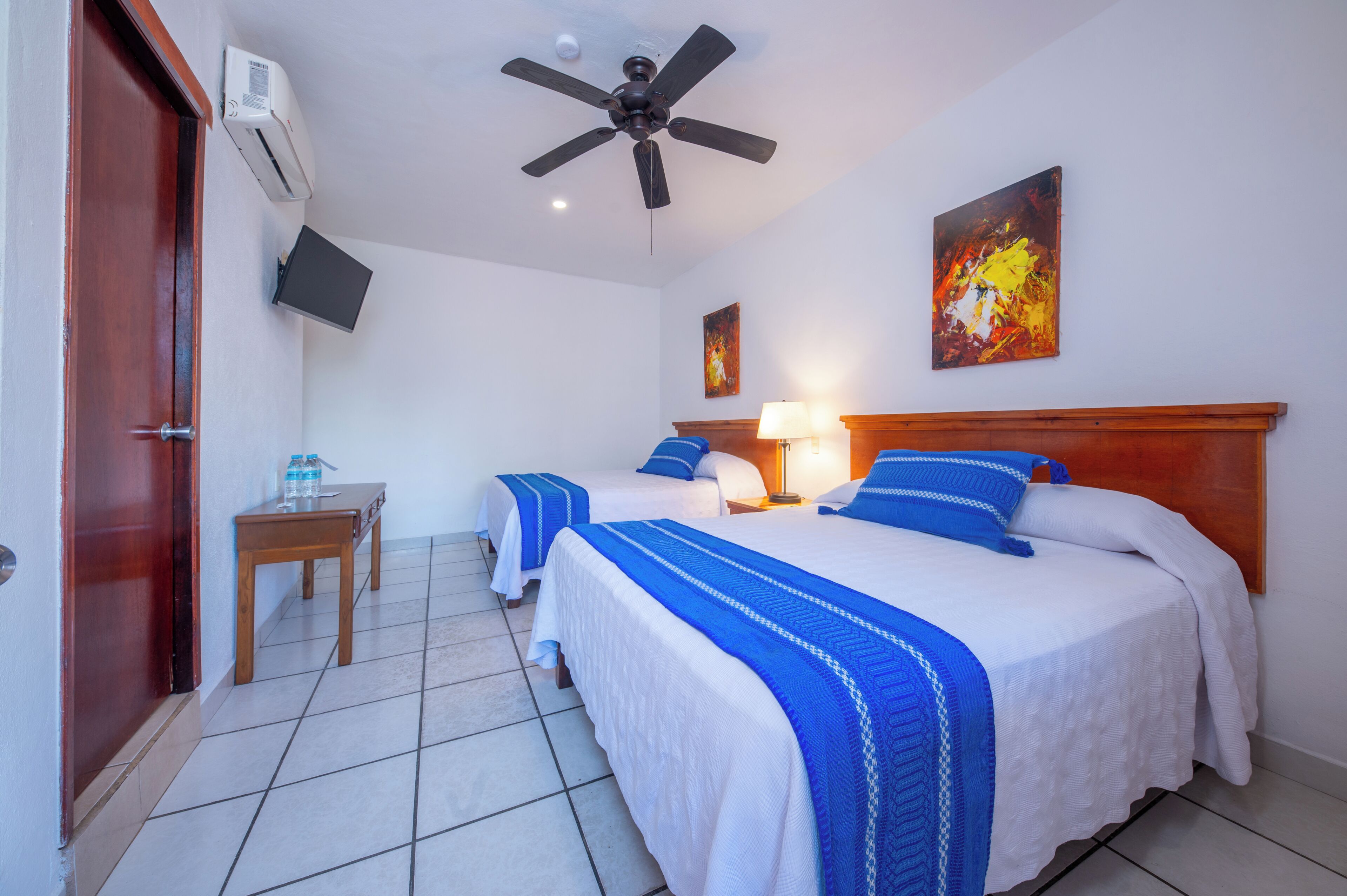 A hotel room with two beds, a ceiling fan, a TV mounted on the wall, and a wooden door.