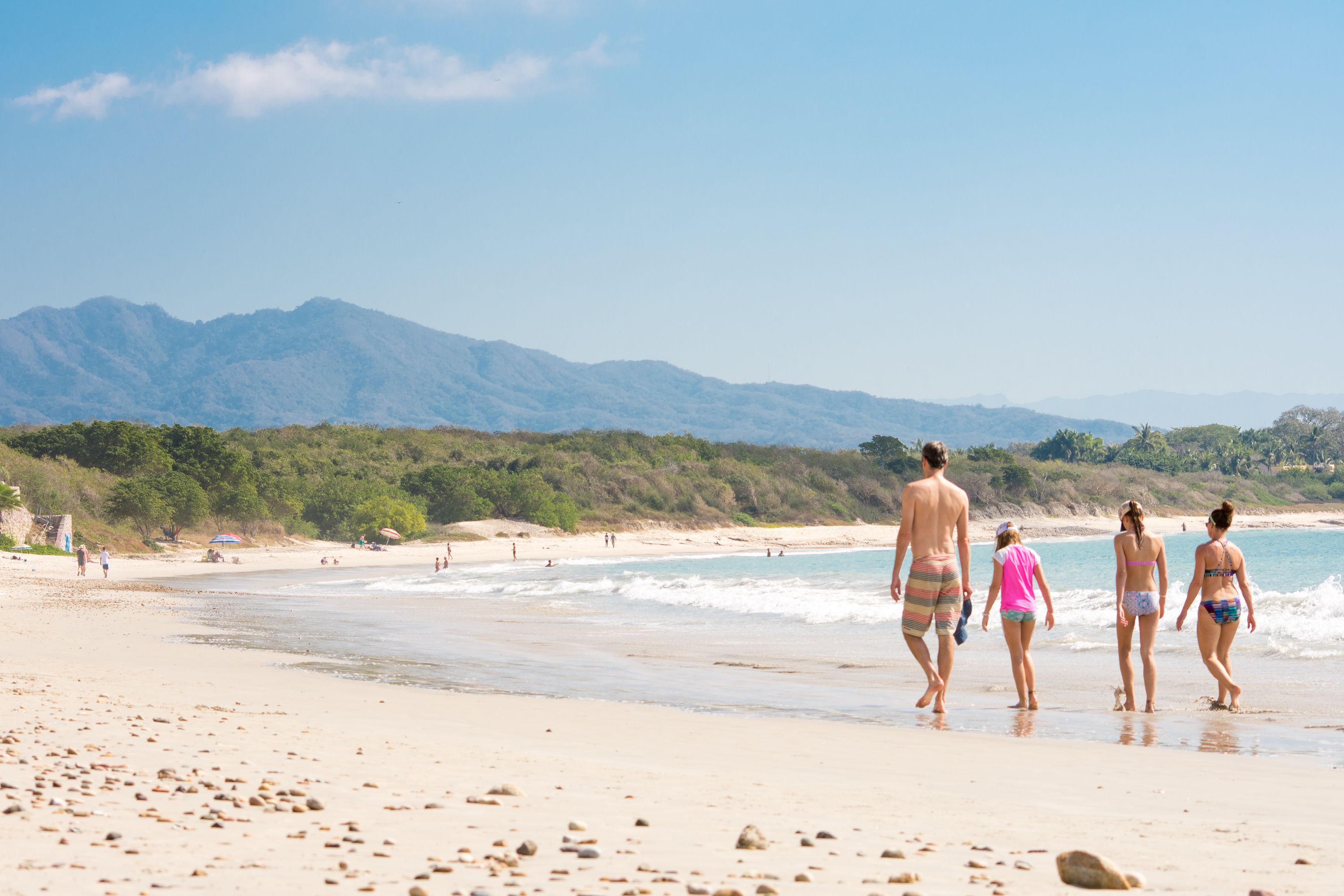 Una playa con gente caminando por la orilla, montañas al fondo y un cielo despejado.