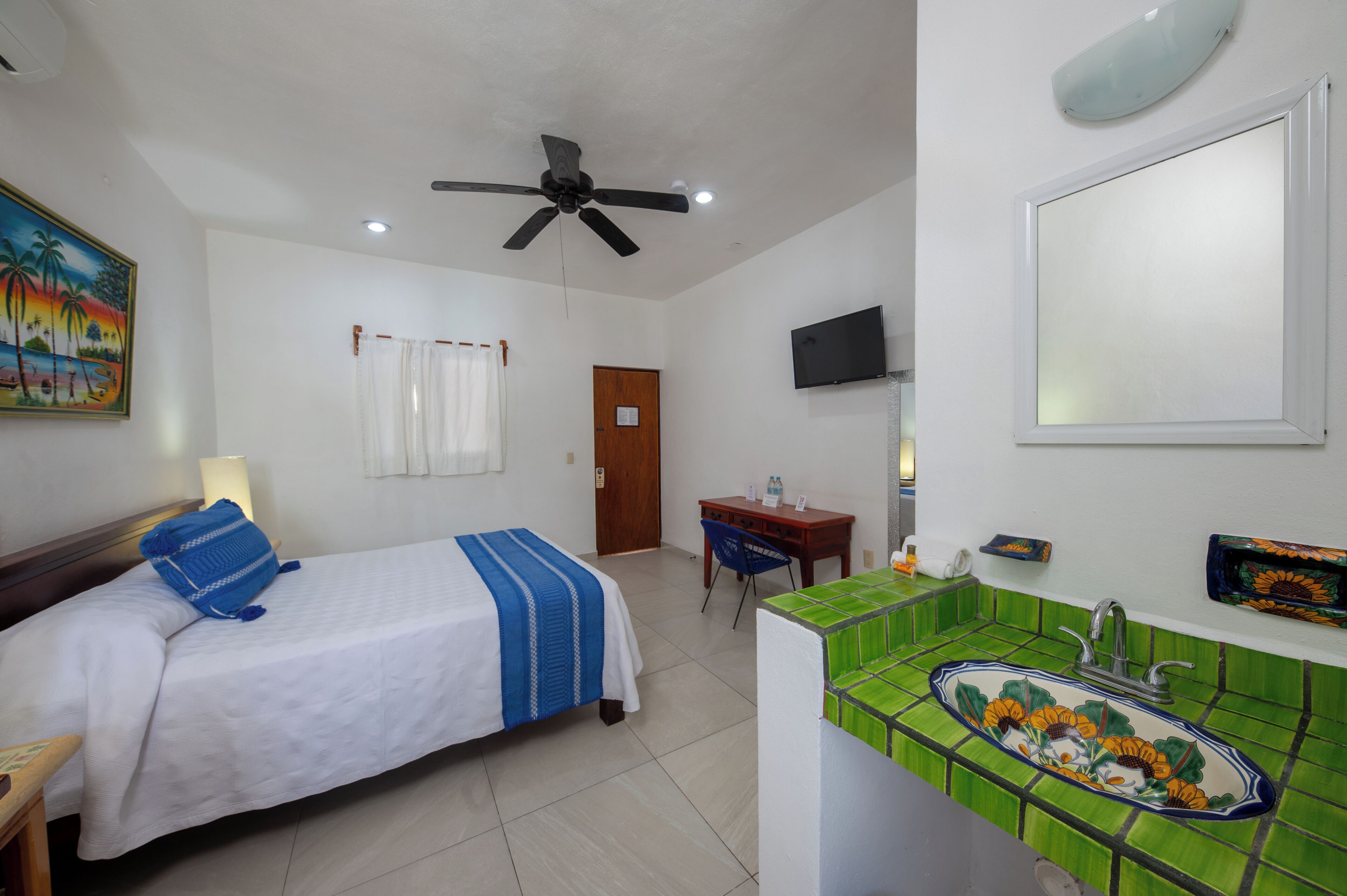 Habitación de hotel con cama, escritorio, silla, ventilador de techo, televisión y baño con lavabo y espejo.
