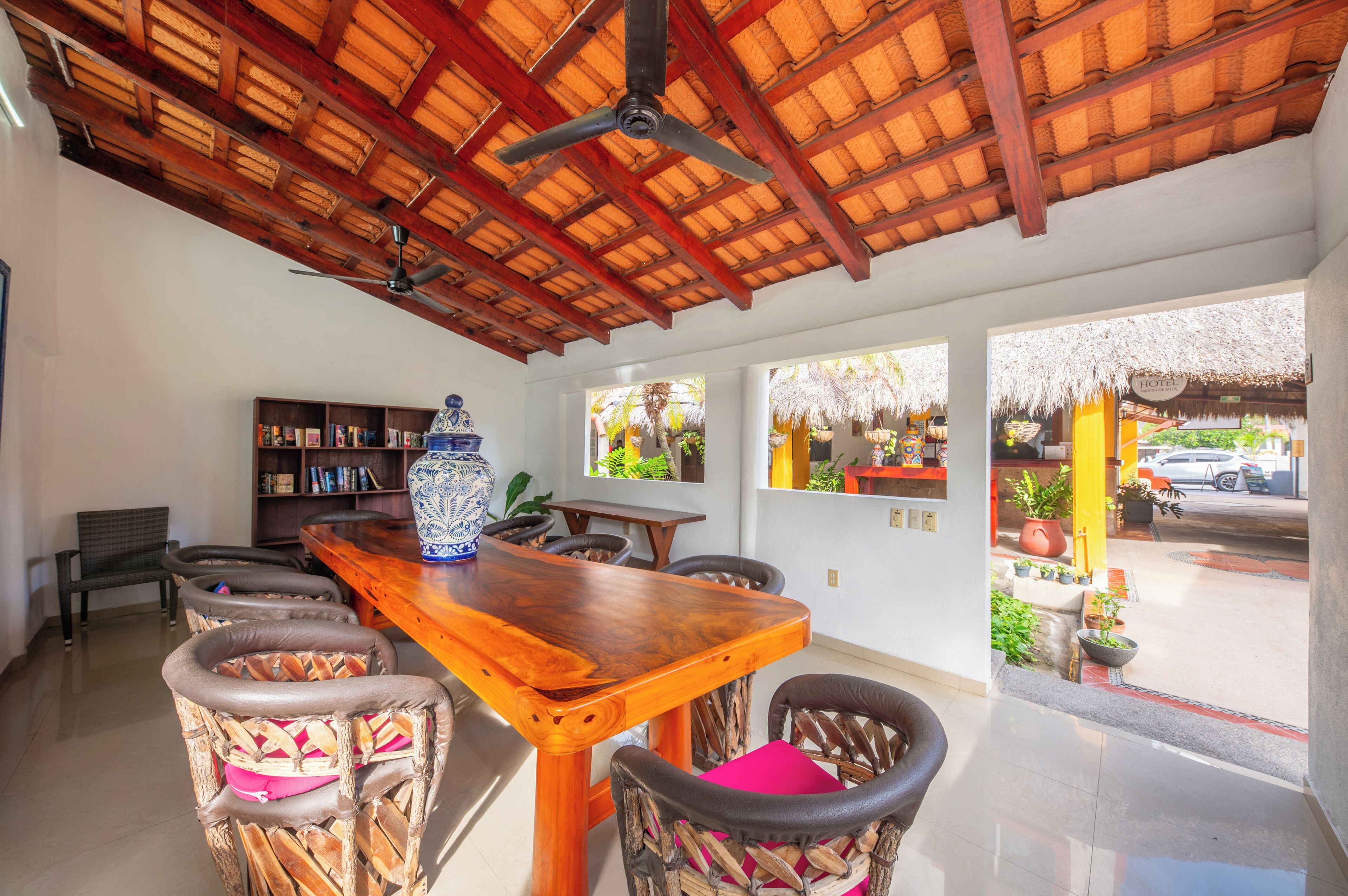 Un comedor con una mesa de madera, sillas de mimbre, ventiladores de techo y vistas a la calle.