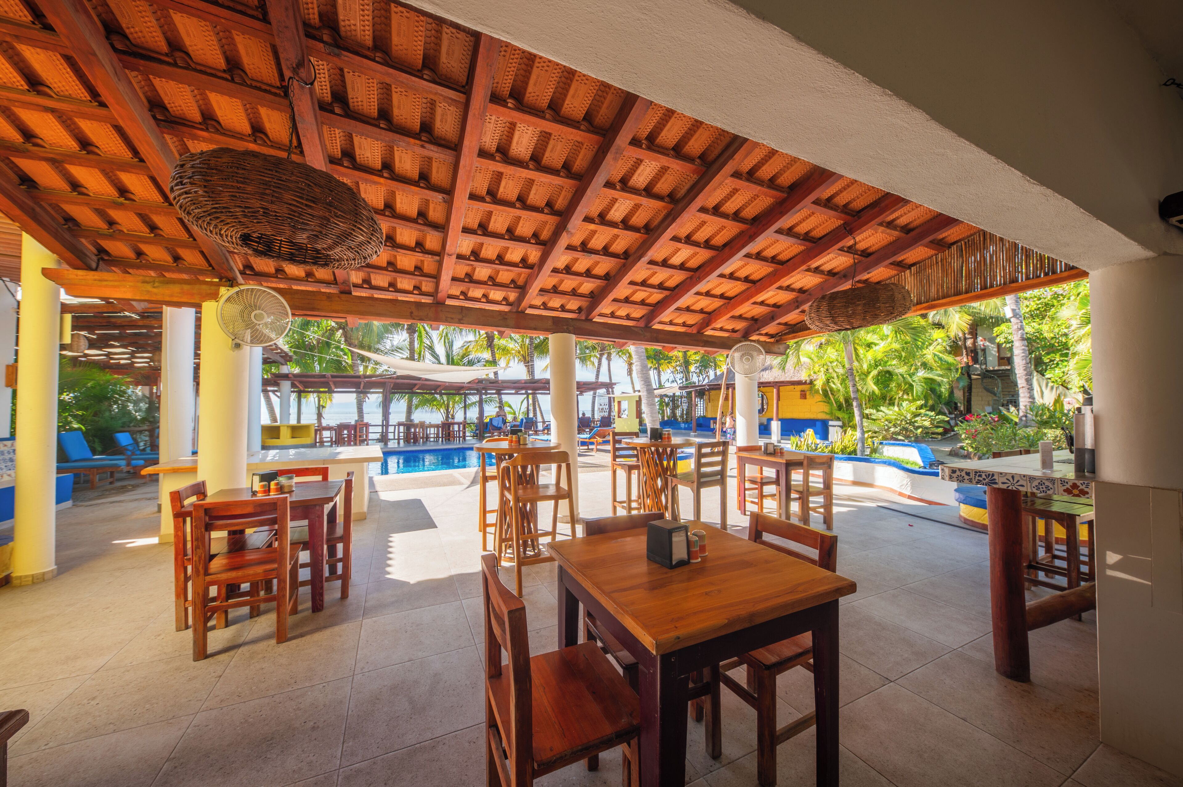 An outdoor dining area with wooden tables and chairs, a pool, and a covered section with a woven ceiling.