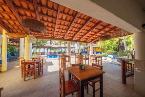 Un área de comedor al aire libre con mesas y sillas de madera, una piscina y una sección cubierta con techo de tejido.