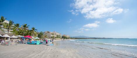 A beach with umbrellas, palm trees, and a building in the background.