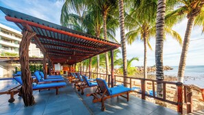A beachside lounge with wooden chairs and a covered area, palm trees, and a view of the ocean. - Hotel Meson de Mita (Punta de Mita)