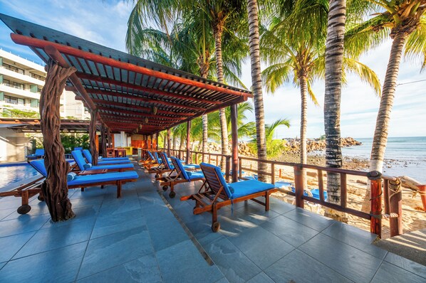 A beachside lounge with wooden chairs and a covered area, palm trees, and a view of the ocean.