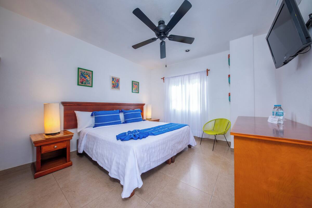 A hotel room with a bed, a desk, a chair, a ceiling fan, and a television.