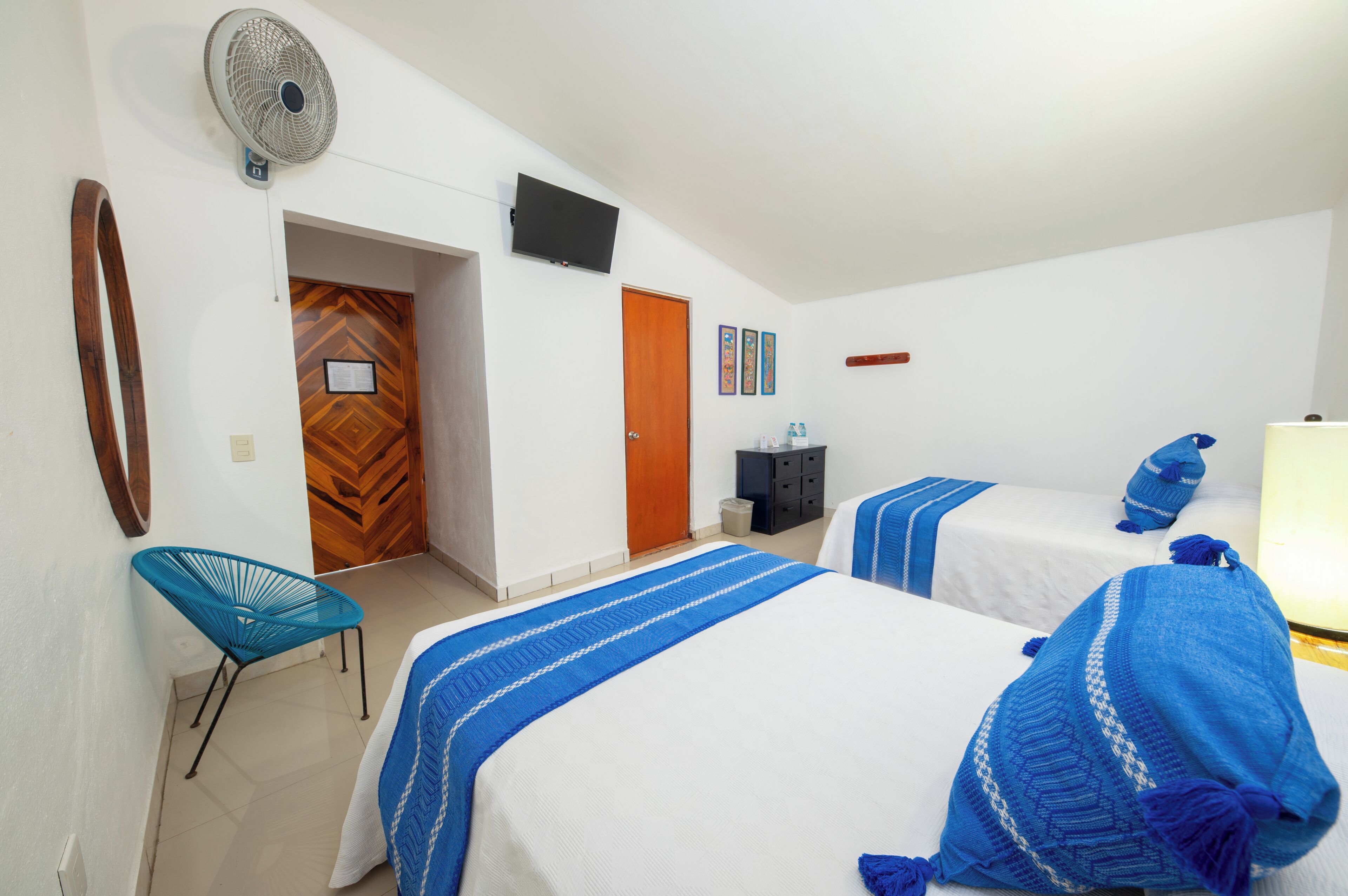 A hotel room with two beds, a TV, a fan, a wooden door, and a blue chair.