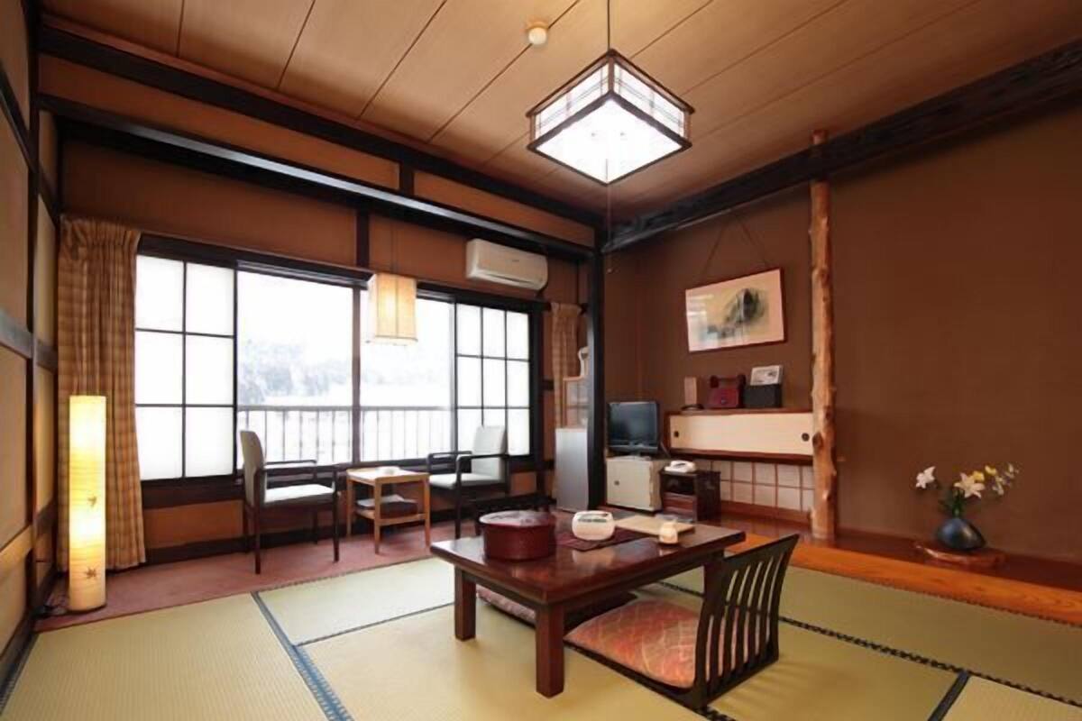 Ein traditioneller japanischer Raum mit Tatami-Boden, einem niedrigen Tisch, Stühlen, einem Fernseher und einem großen Fenster.