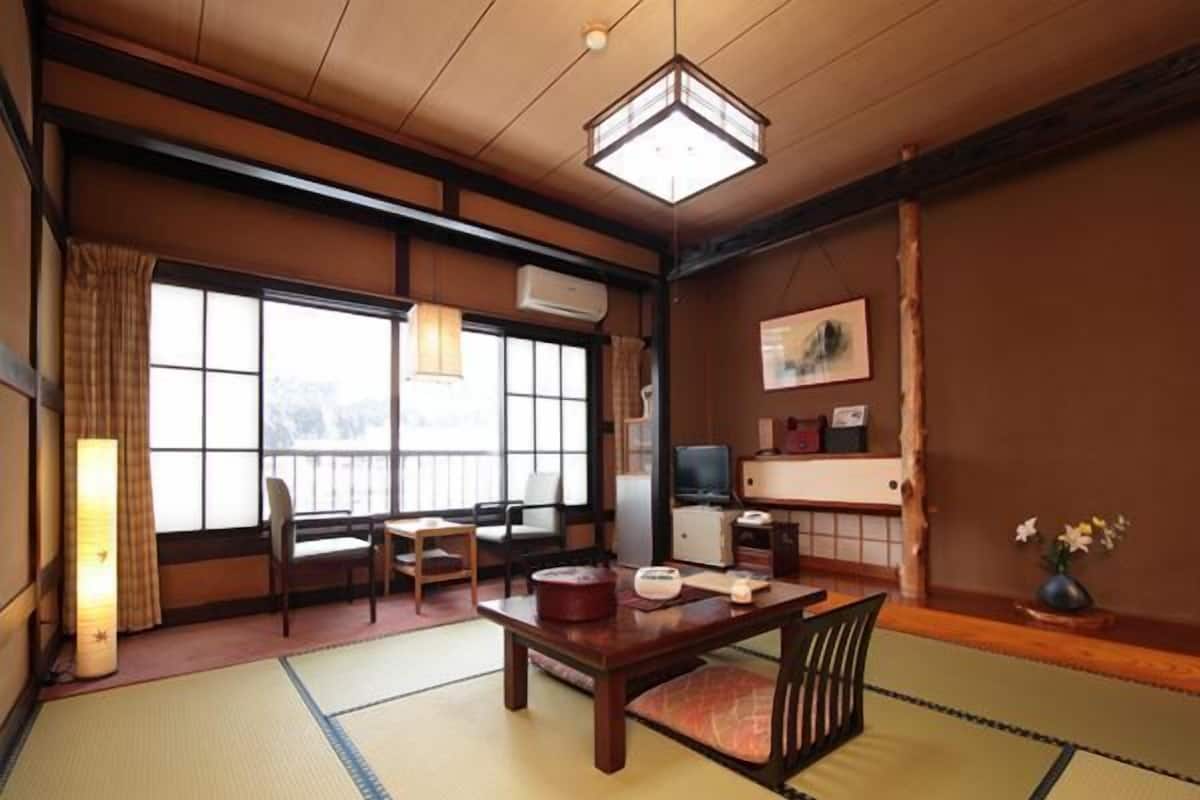 Una habitación japonesa tradicional con suelo de tatami, una mesa baja, sillas, un televisor y un amplio ventanal.
