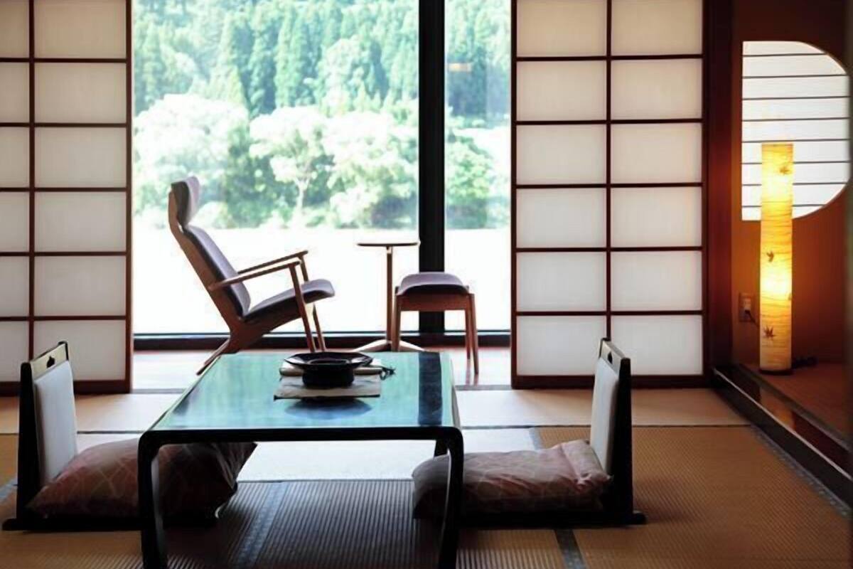 Ein traditioneller japanischer Raum mit Tatami-Böden, einem niedrigen Tisch, einem Stuhl und Blick auf einen Wald durch große Fenster.