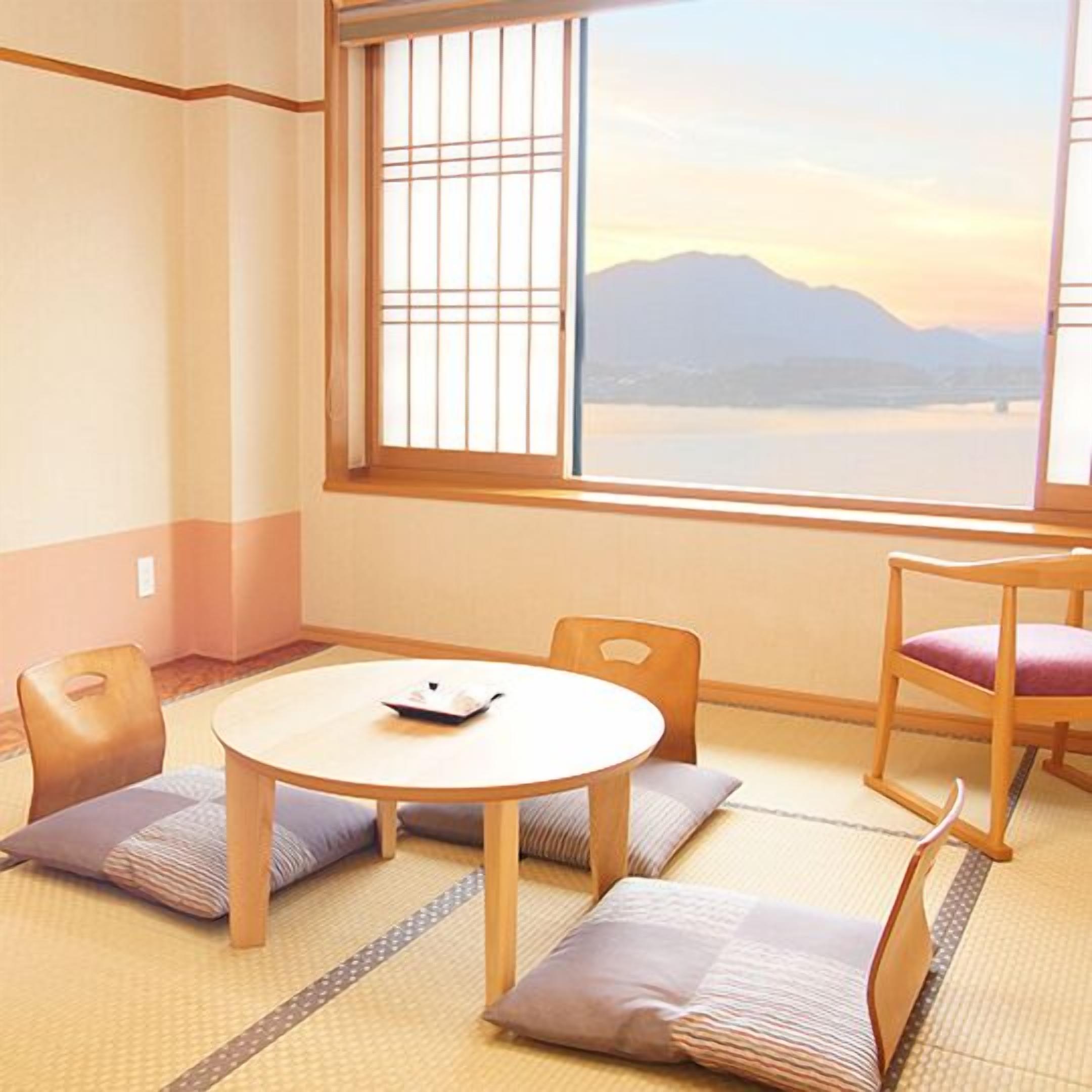 Een kamer met tatami vloerbedekking, een ronde tafel, twee stoelen en uitzicht op bergen door een raam met een rasterpatroon.