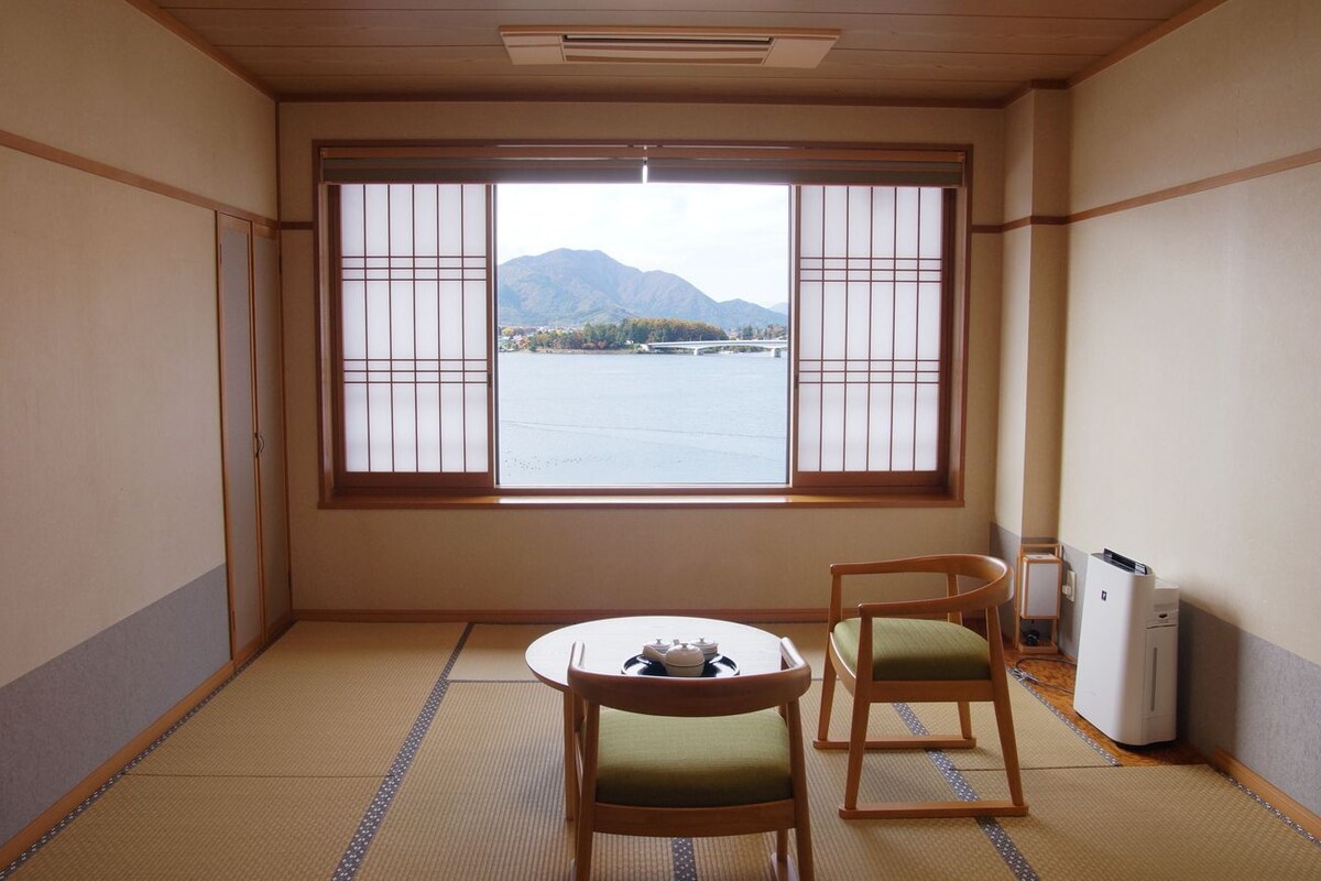 A room with tatami flooring, a traditional Japanese sliding door, a table with a tea set, two chairs, and a view of a mountain and water.