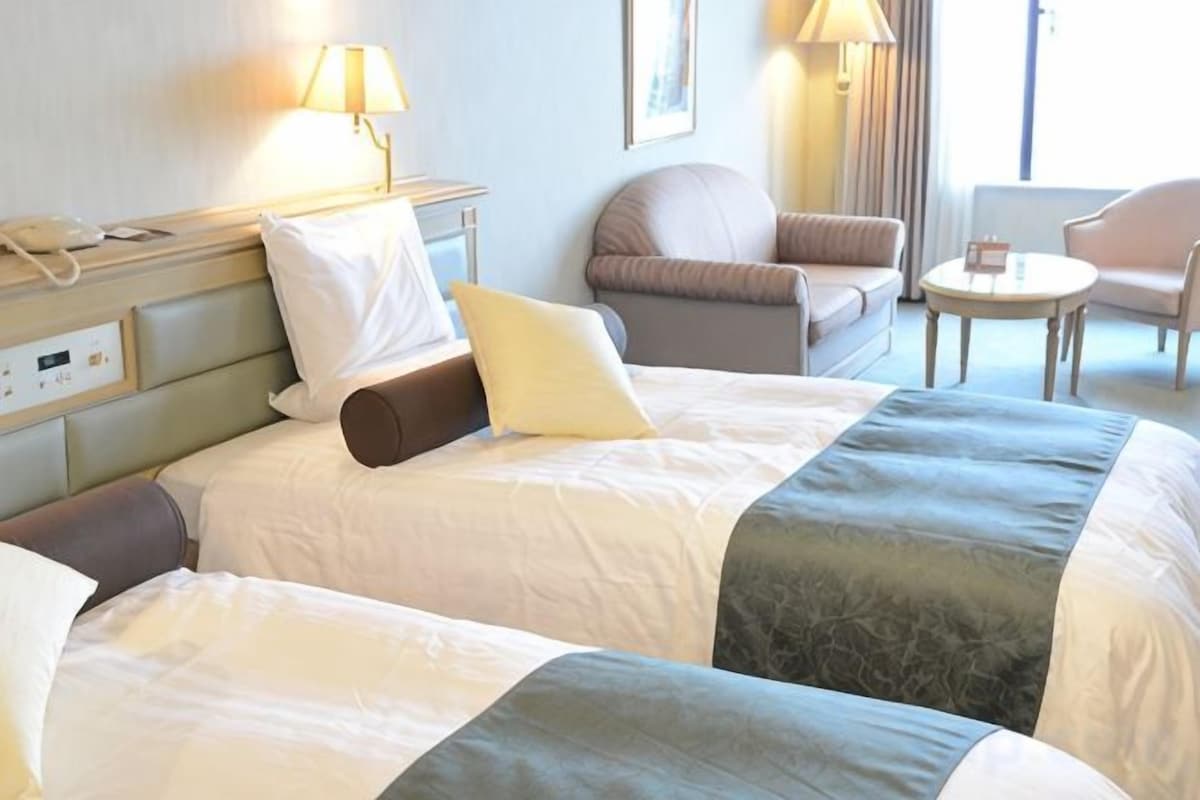 Et hotelværelse med to senge, et sengebord med lampe, en stol og et lille rundt bord.