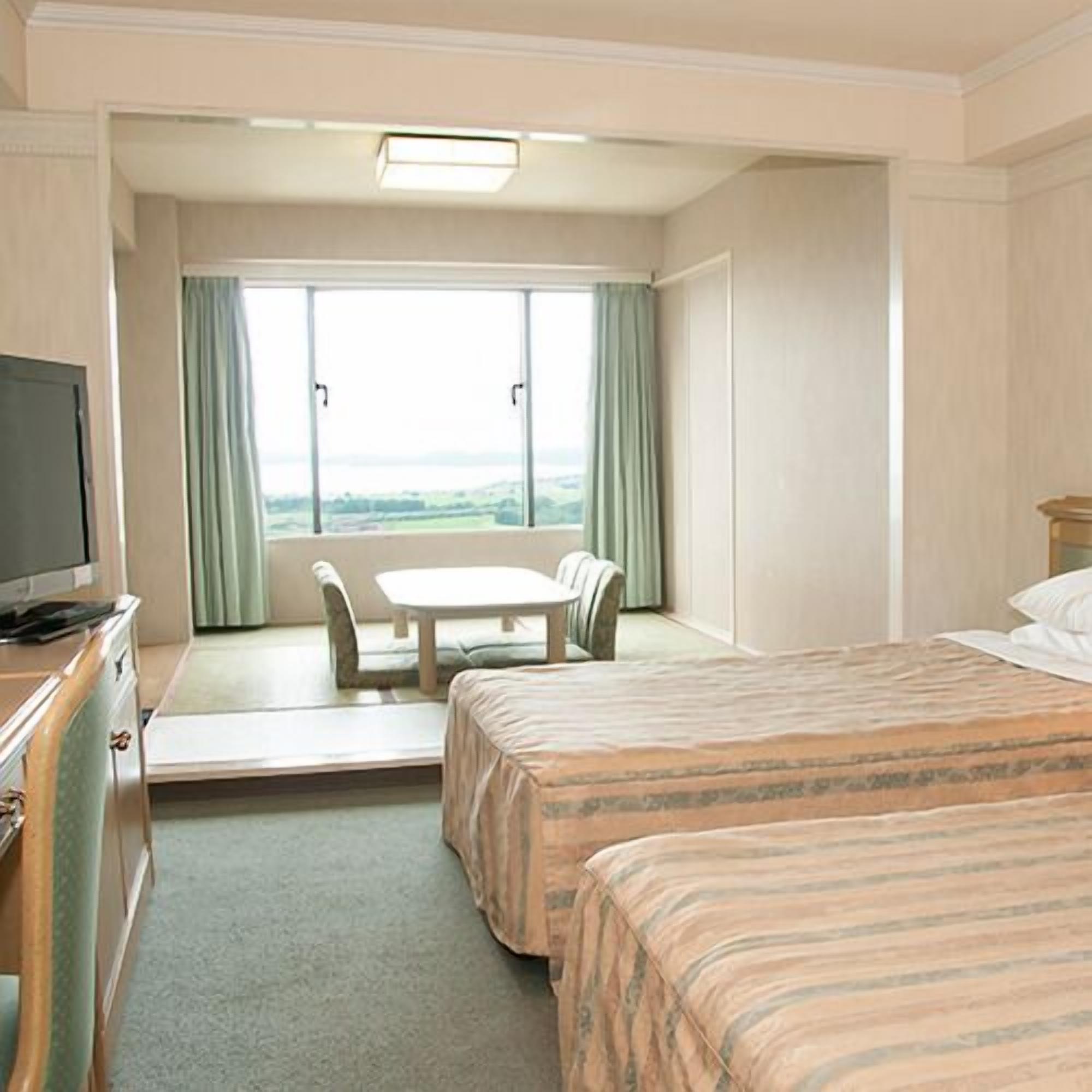 A hotel room with a bed, a desk with a chair, a television, and a large window with curtains.