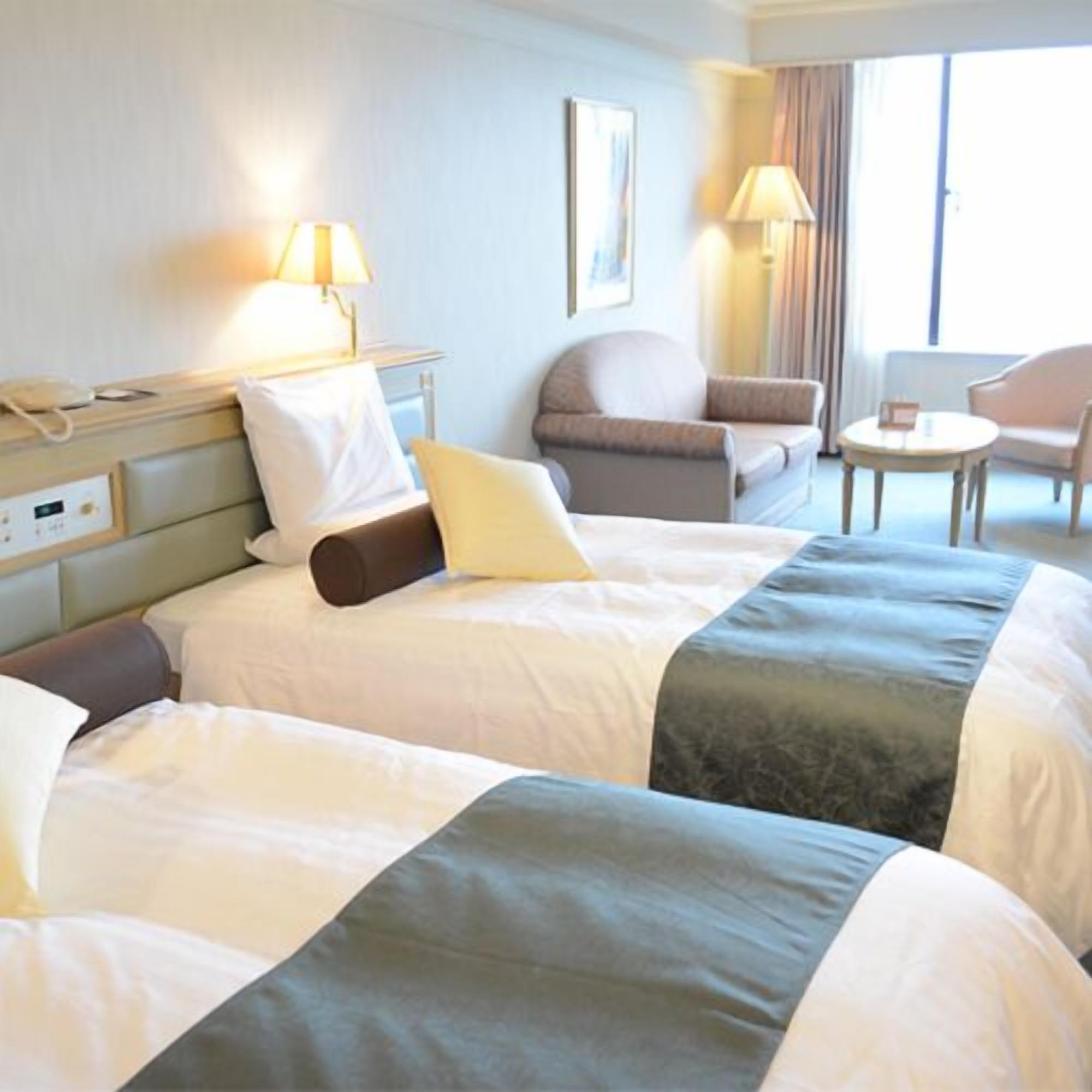 A hotel room with two beds, a bedside table with a lamp, a chair, a small table, and a window with curtains.