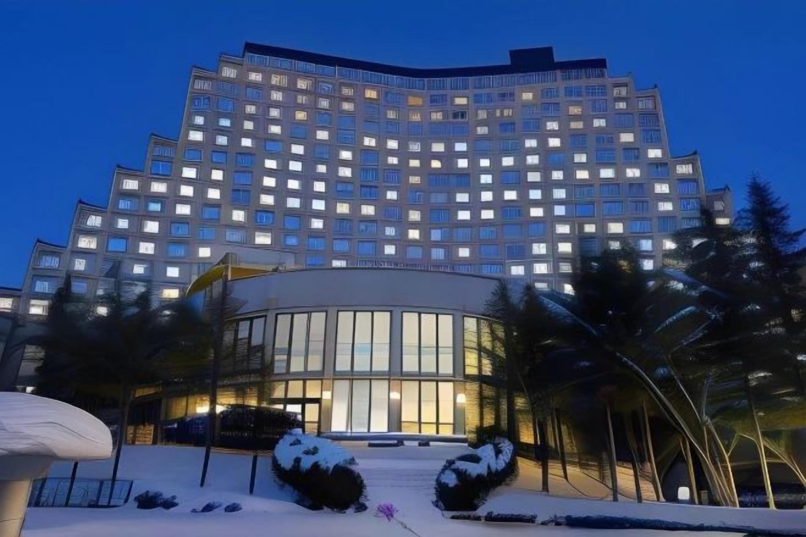 Een hotel met meerdere verdiepingen, voorzien van een glazen gevel in het midden, omringd door aangelegde zones met vormgesnoeide hagen.