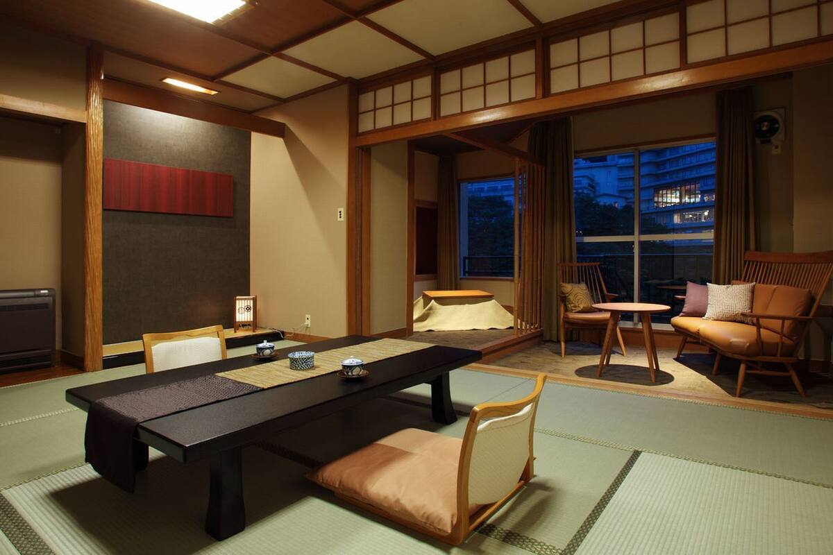 Une pièce de style japonais traditionnel, avec un sol recouvert de tatamis, une table basse, des chaises et une vue sur la ville la nuit.