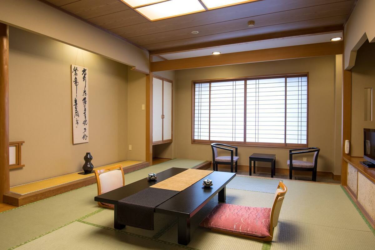 Une pièce de style japonais traditionnel, avec un sol recouvert de tatamis, une table basse, des chaises, une porte coulissante et une grande fenêtre.