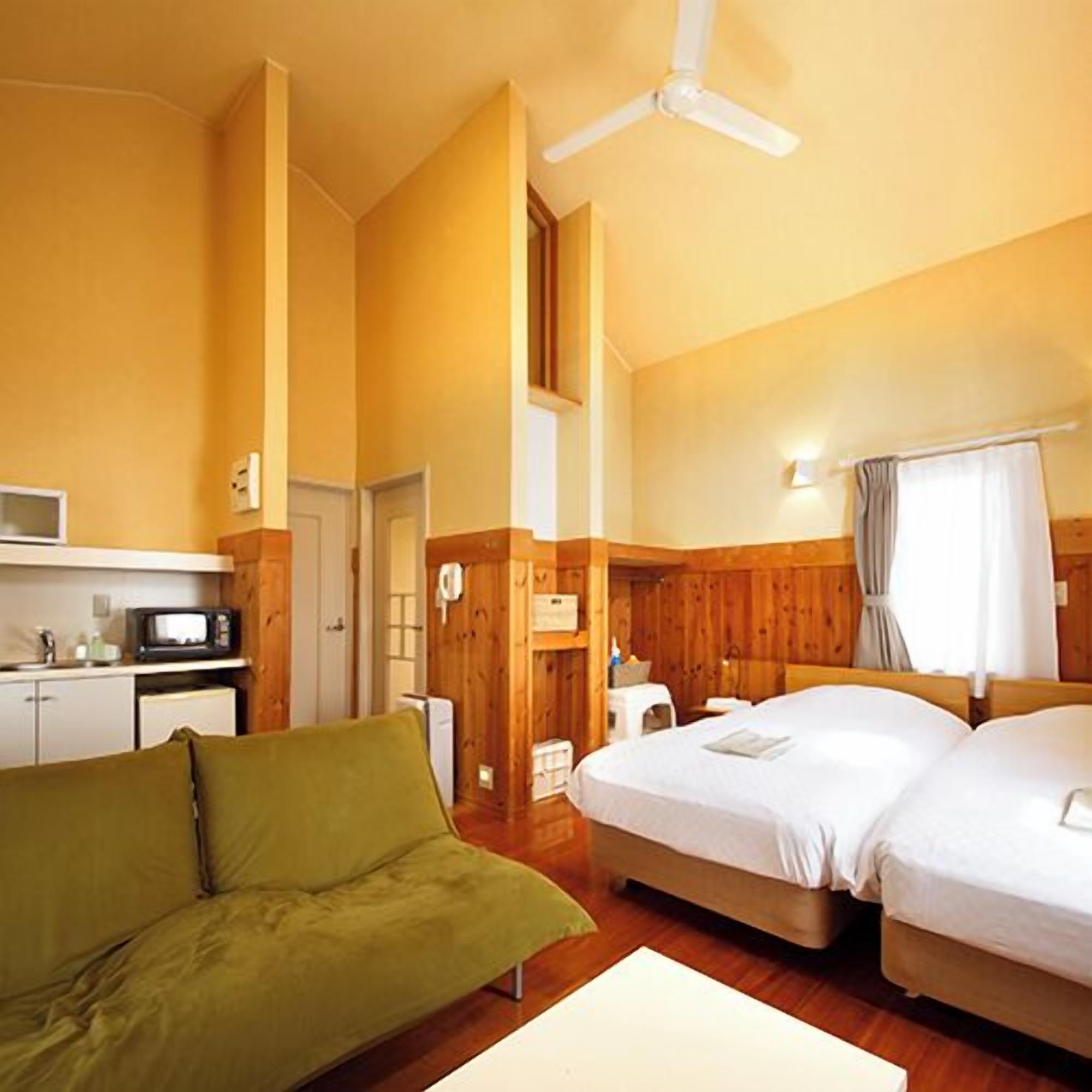 A room with a green sofa, two beds, a ceiling fan, a microwave, and wooden walls.