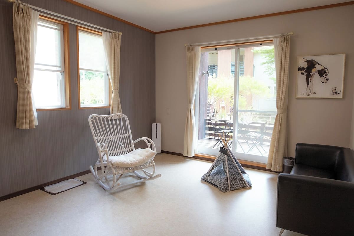 Habitació amb una cadira bressol blanca, un butac armchair negre, una taula petita i una porta corredissa de vidre que dona a una zona exterior.