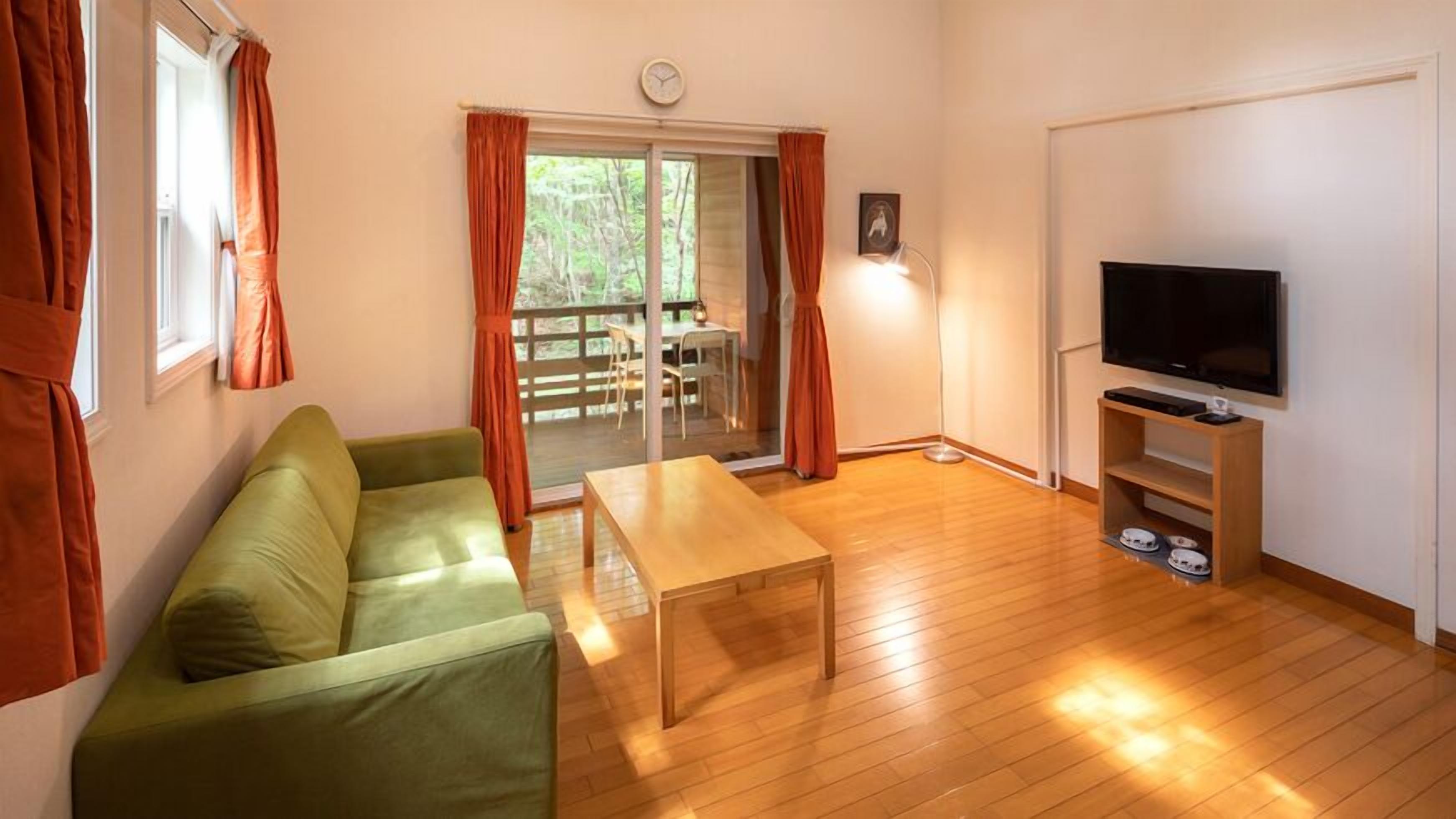 A living room with a green sofa, a wooden table, a TV on a stand, and a sliding glass door leading to an outdoor area.