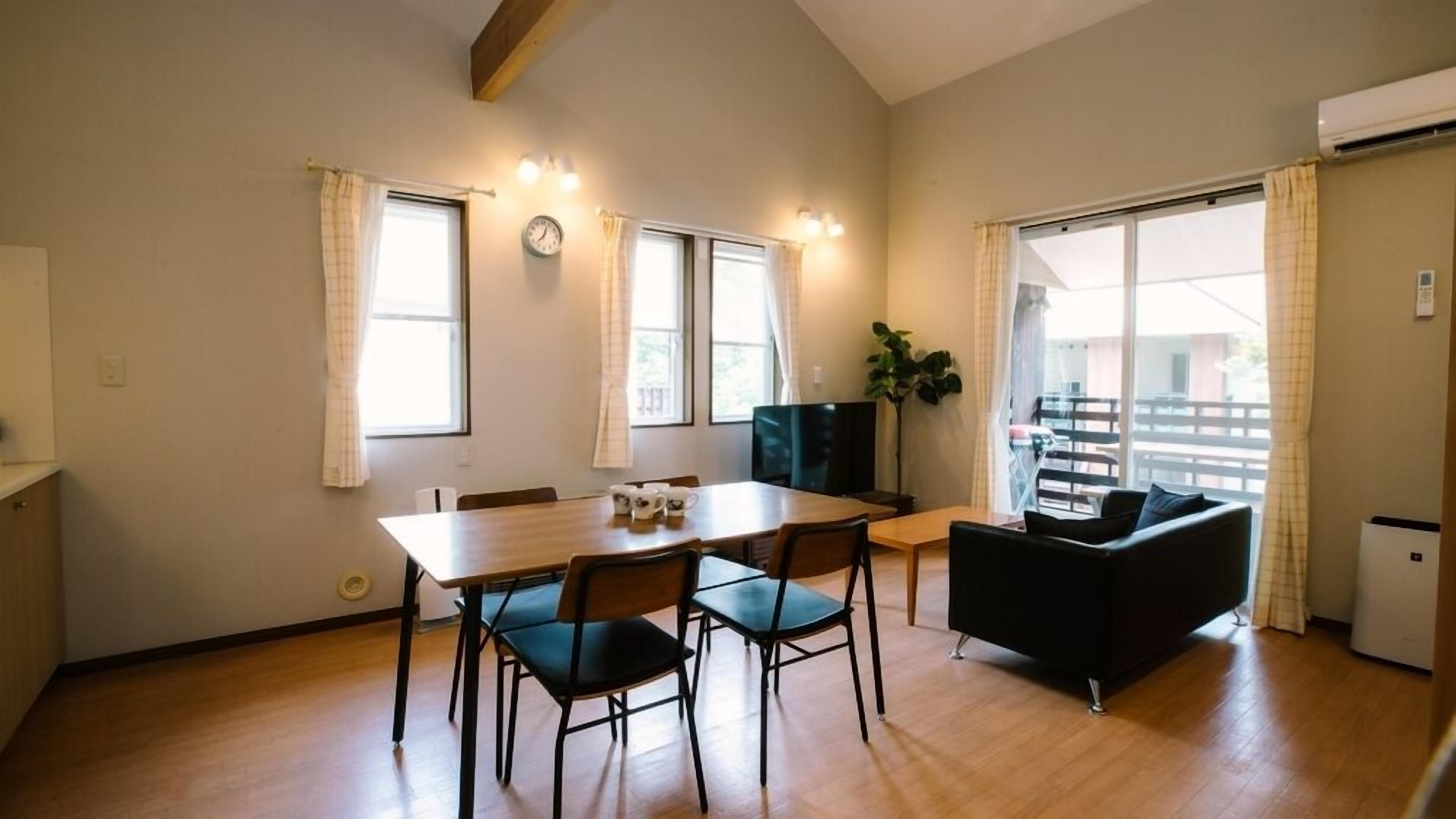 A room with a dining table, chairs, a sofa, a TV, and a balcony with a railing.