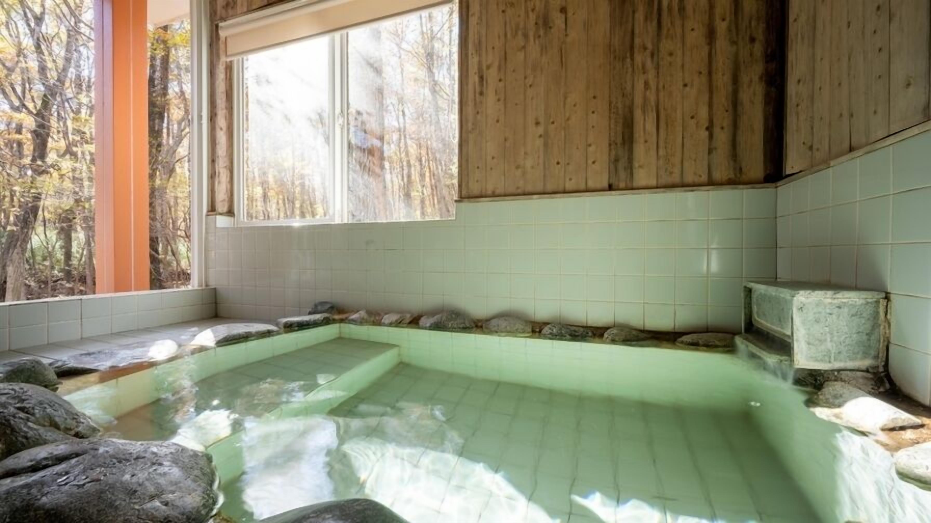 A hot spring bath with tiled walls and wooden beams, surrounded by rocks and a window overlooking trees.
