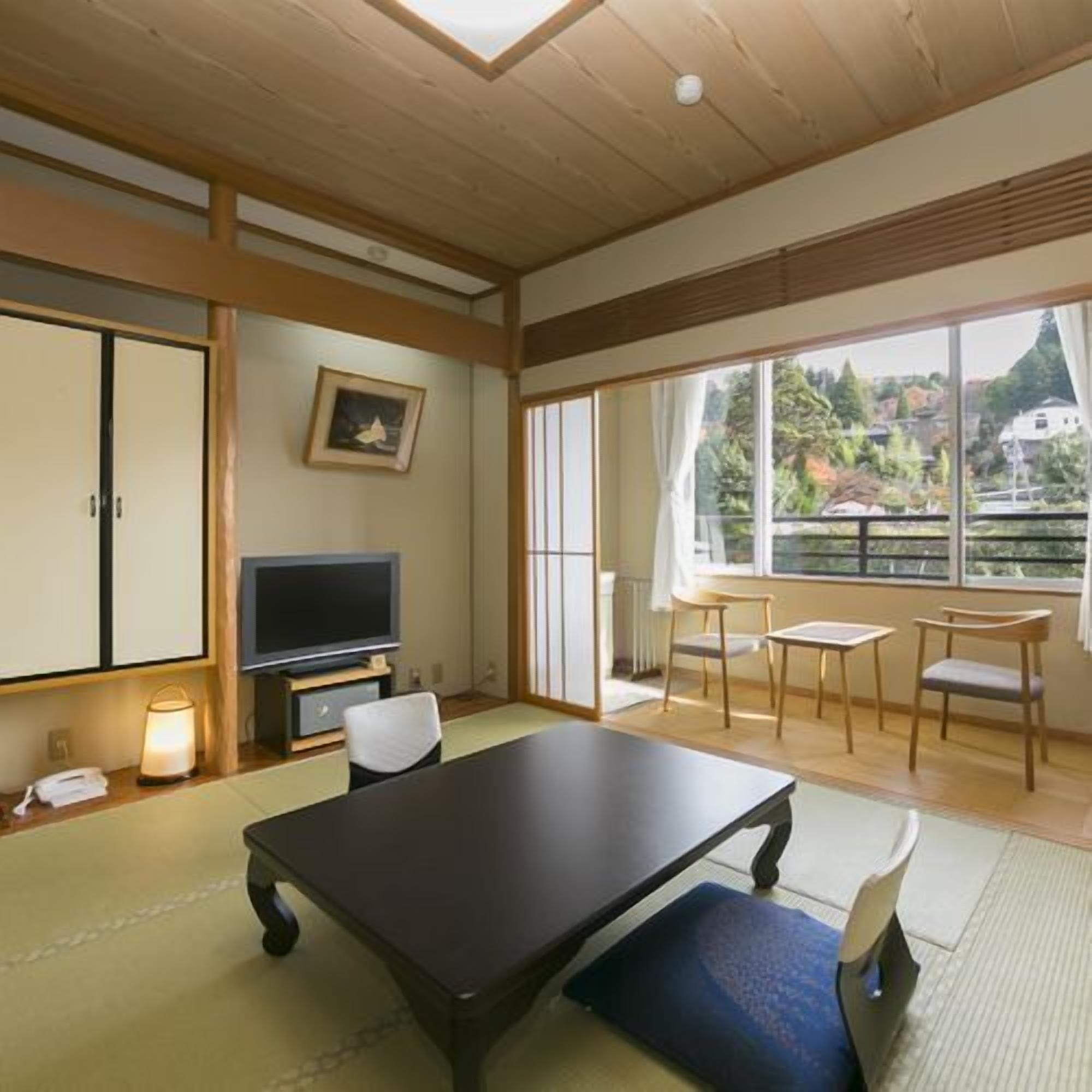 A room with a TV, a desk, chairs, a picture on the wall, and a view through large windows.