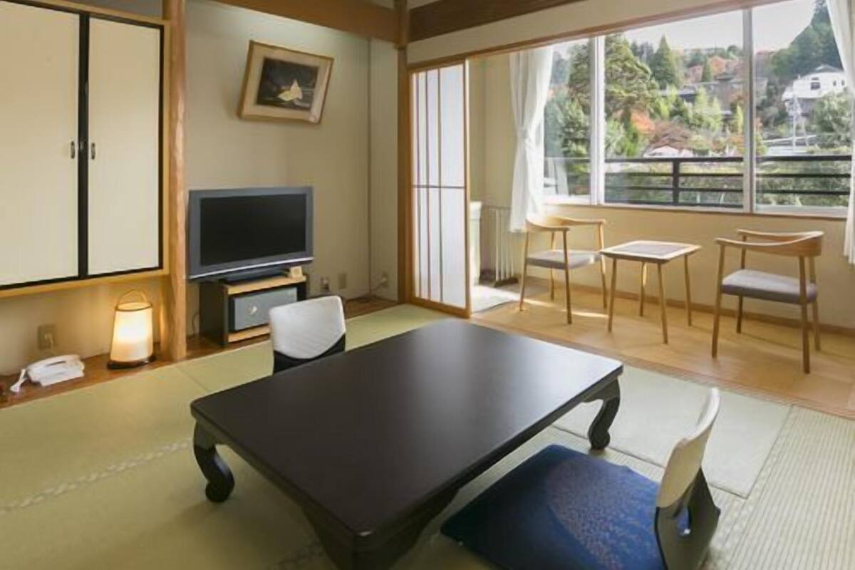 A room with a TV, a desk, chairs, a picture on the wall, and large windows.