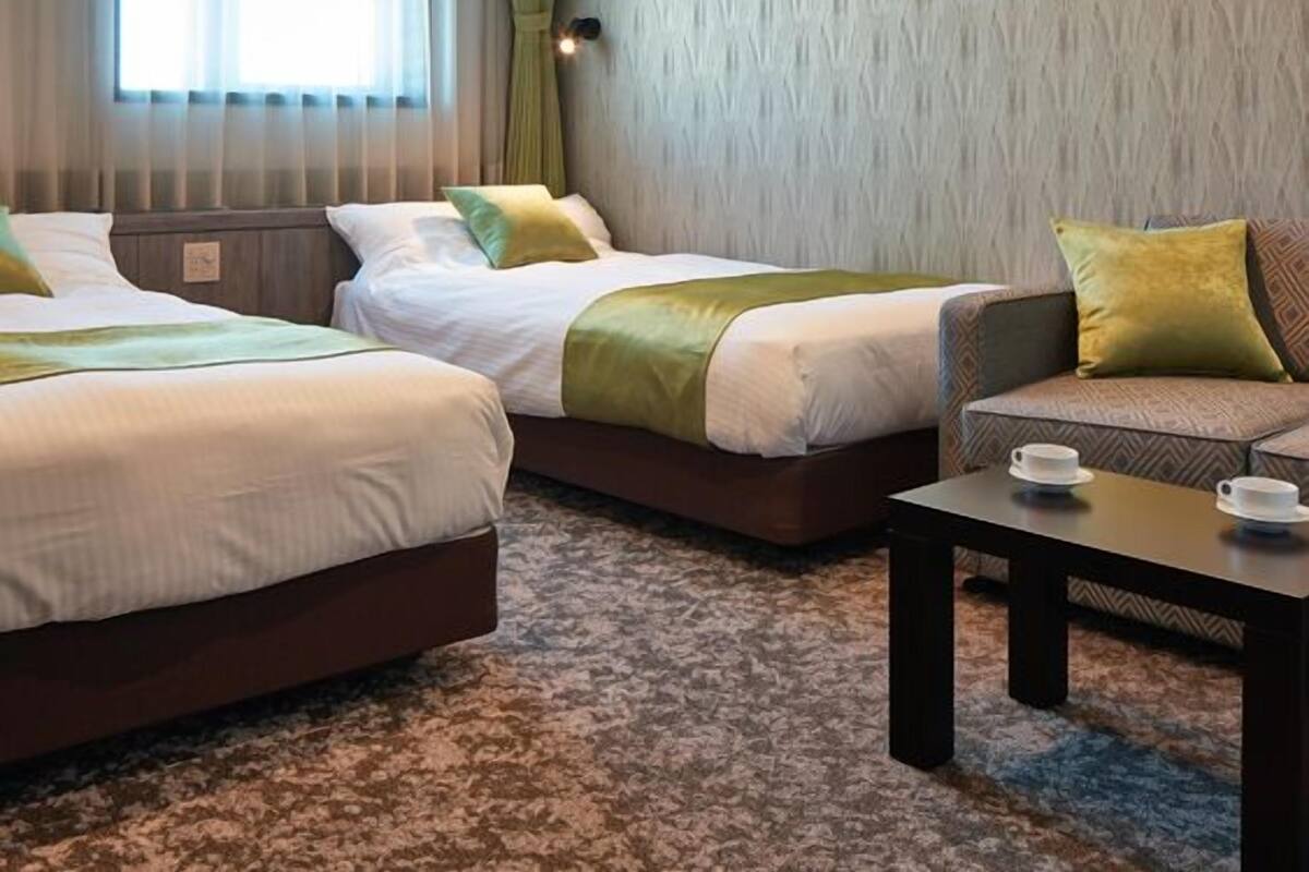 Habitación de hotel con dos camas, un sofá, una mesita con tazas y una ventana con cortinas.