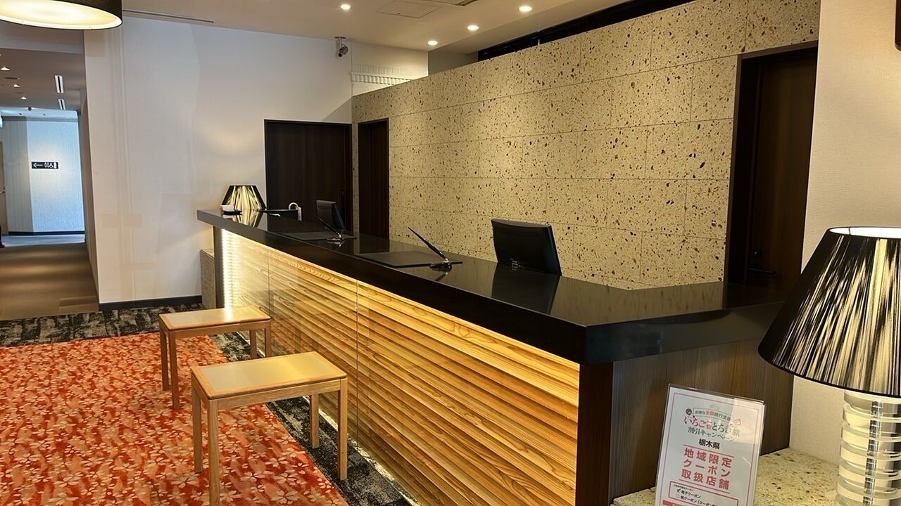 Un espace d’accueil d’hôtel avec un comptoir en bois, deux tabourets, une chaise noire et un panneau avec du texte en japonais.