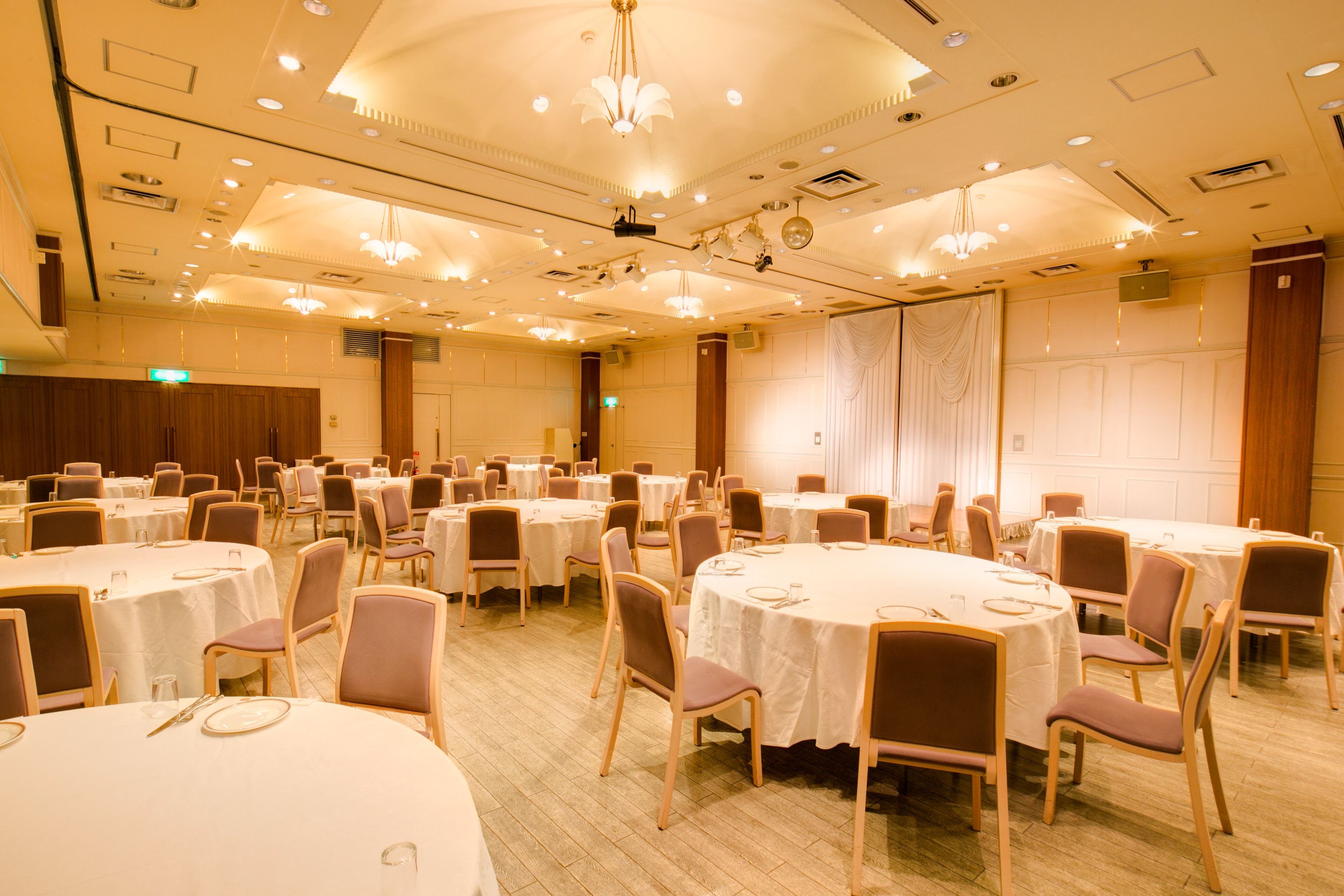 Une salle de banquet avec des tables et des chaises rondes, équipée de luminaires au plafond et d’un système de ventilation.