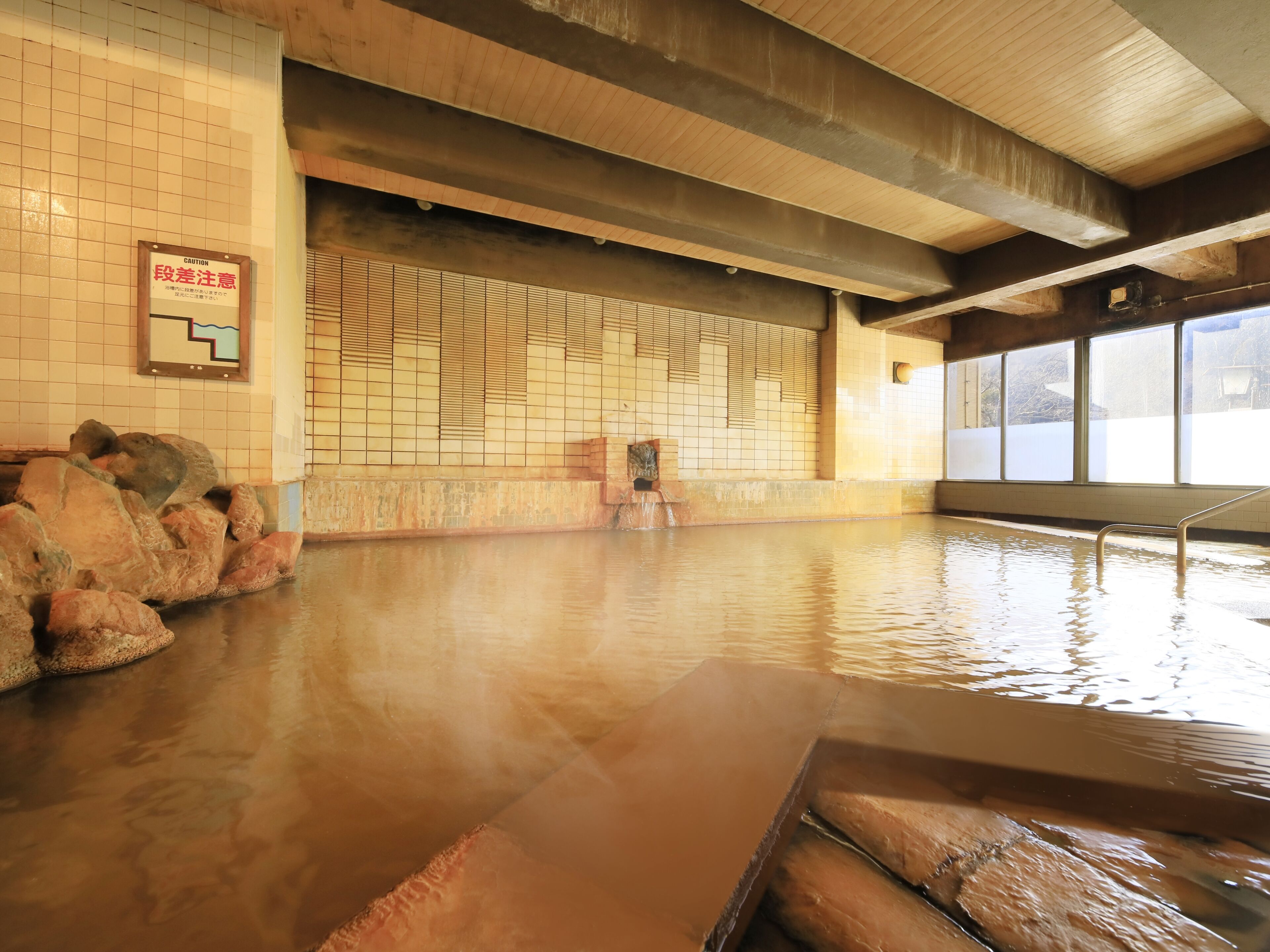A hot spring bath with tiled walls, a wooden ceiling, and a caution sign.
