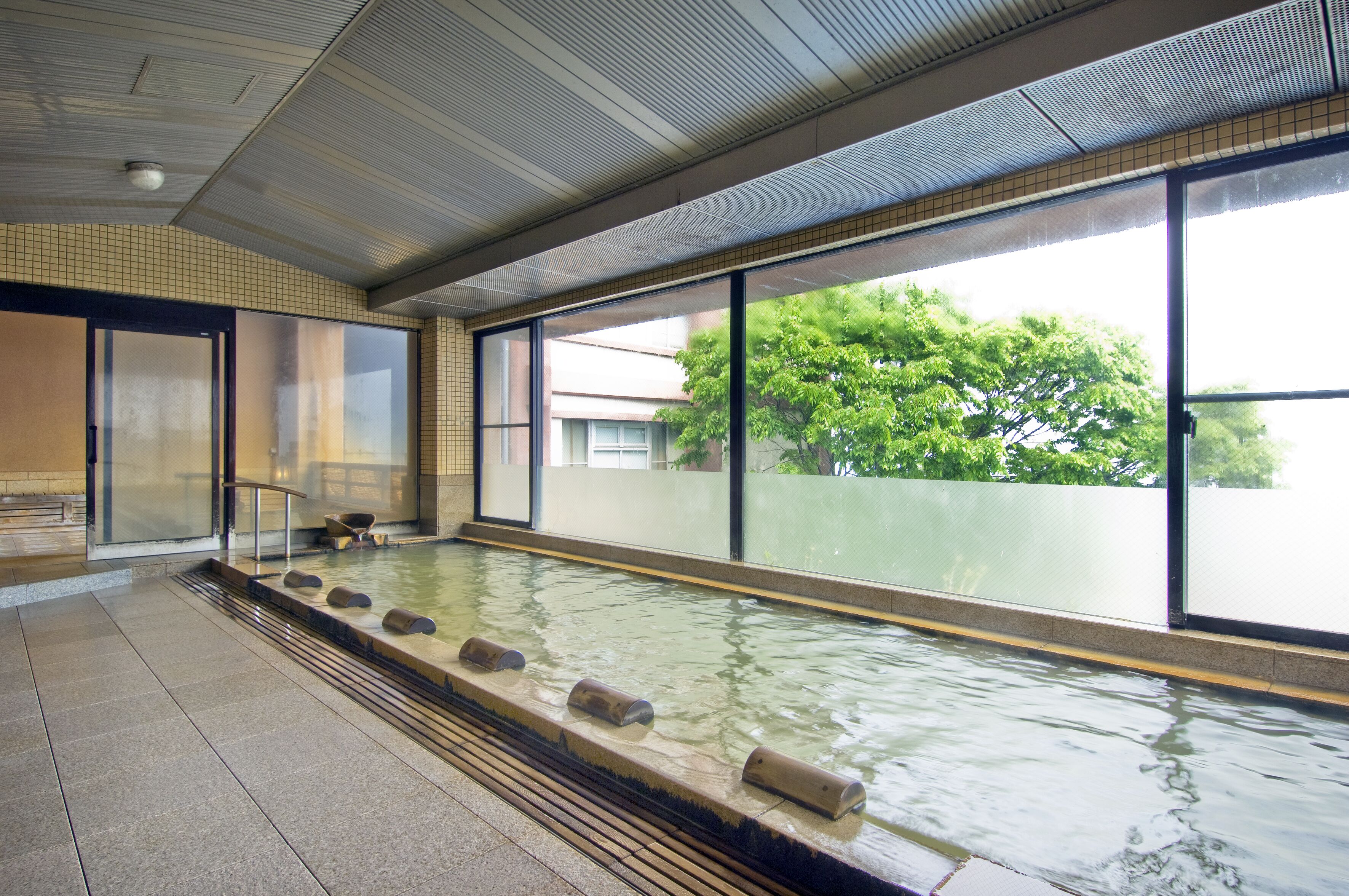 A large indoor pool with clear glass walls, a tiled floor, and a view of trees outside.