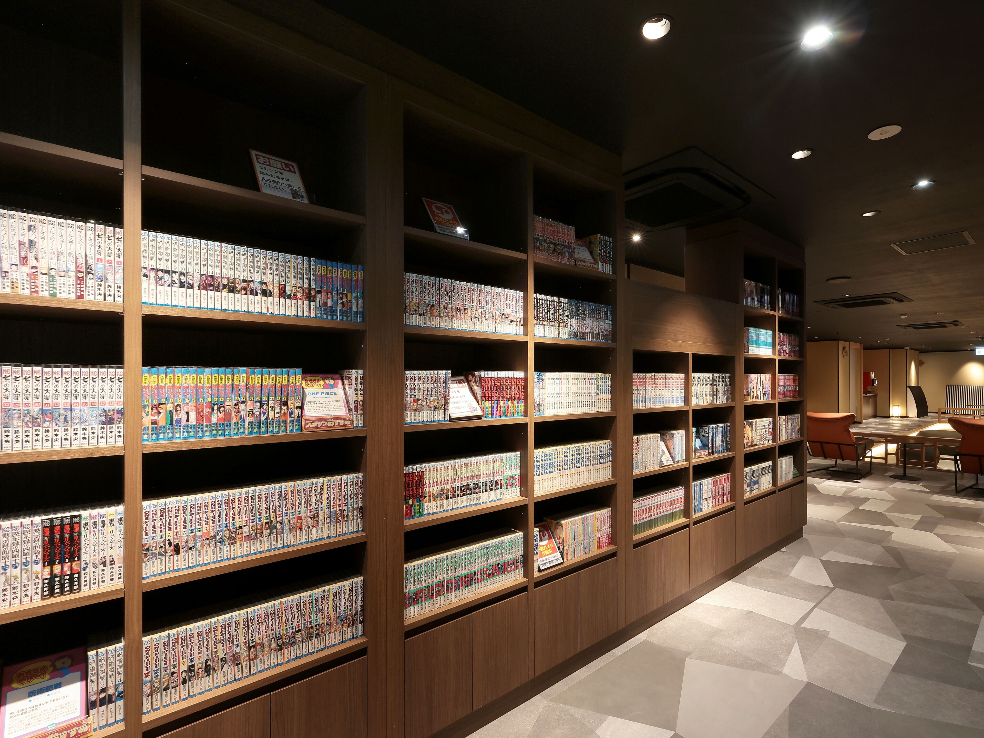 A library with wooden shelves filled with manga books, a table with chairs, and a tiled floor.