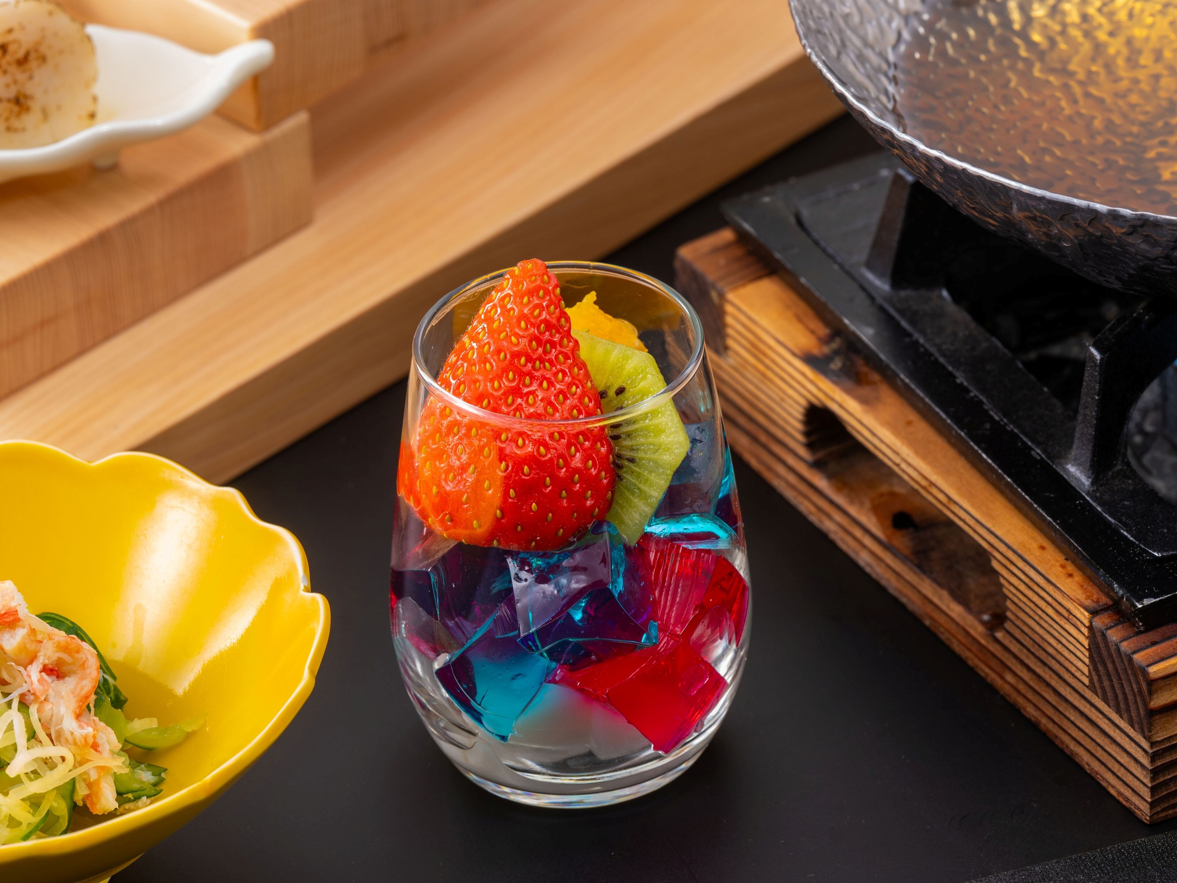 A glass with colorful ice cubes and a strawberry, next to a yellow bowl with food.