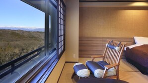 A hotel room with a large window offering a view of a mountainous landscape. There is a wooden chair with a cushion, a bed with a purple bedspread, and a wooden headboard. - Kishigon Ryokan (Shibukawa)